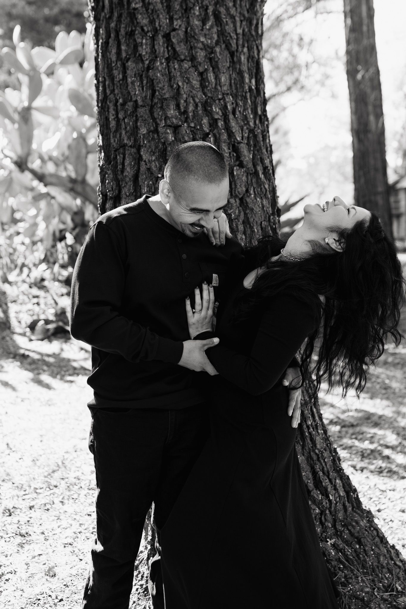 A romantic black and white photo of a couple embracing against a tree trunk in a forest setting.