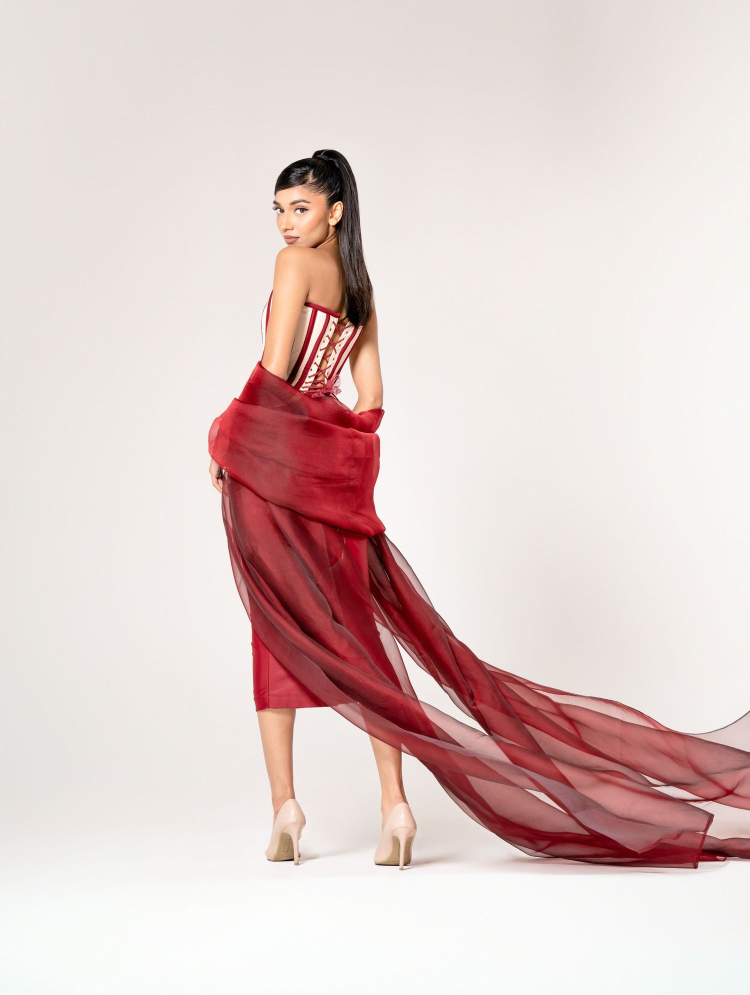 Graceful figure in a stunning red tulle gown with flowing train and beige heels poses against light background.