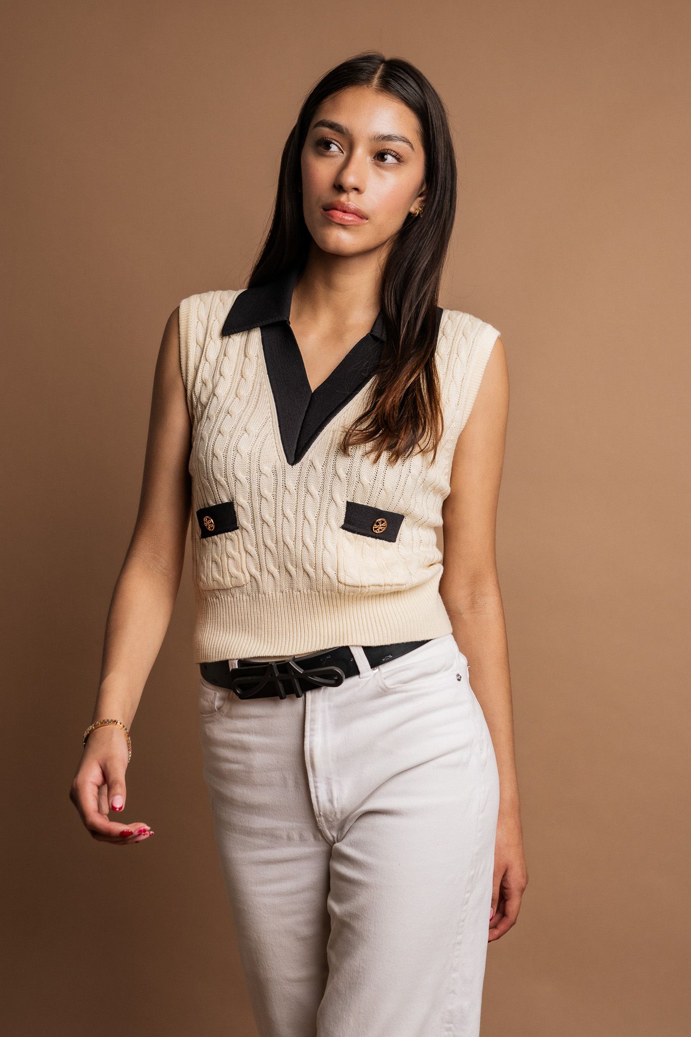 A person in a cream cable knit vest with black collar details poses against a brown backdrop wearing white denim pants.