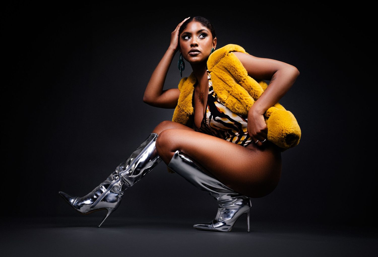 Model wearing yellow outfit and metallic silver boots poses dramatically against dark background.