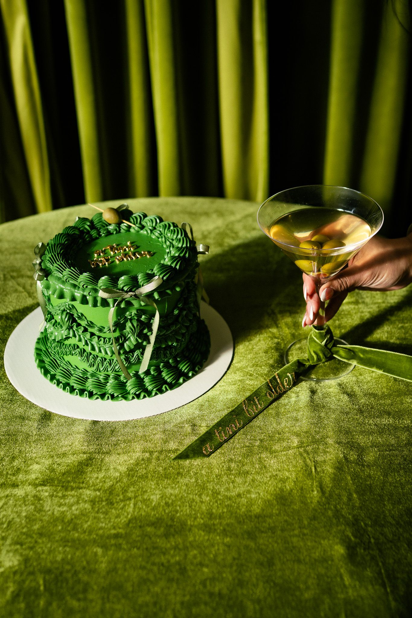 A small green frosted cake with decorative piping sits beside a martini glass on a green velvet tablecloth in moody lighting.
