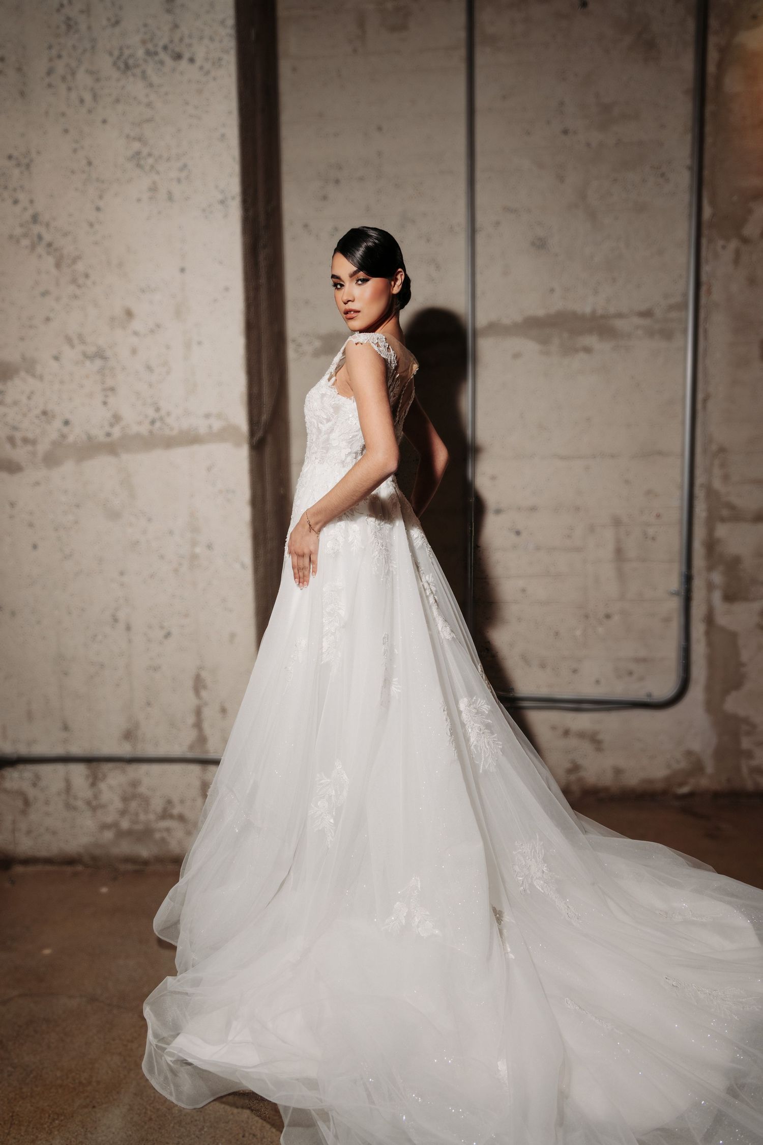 A stunning white wedding gown with a flowing train and detailed lace bodice photographed against a textured concrete wall.