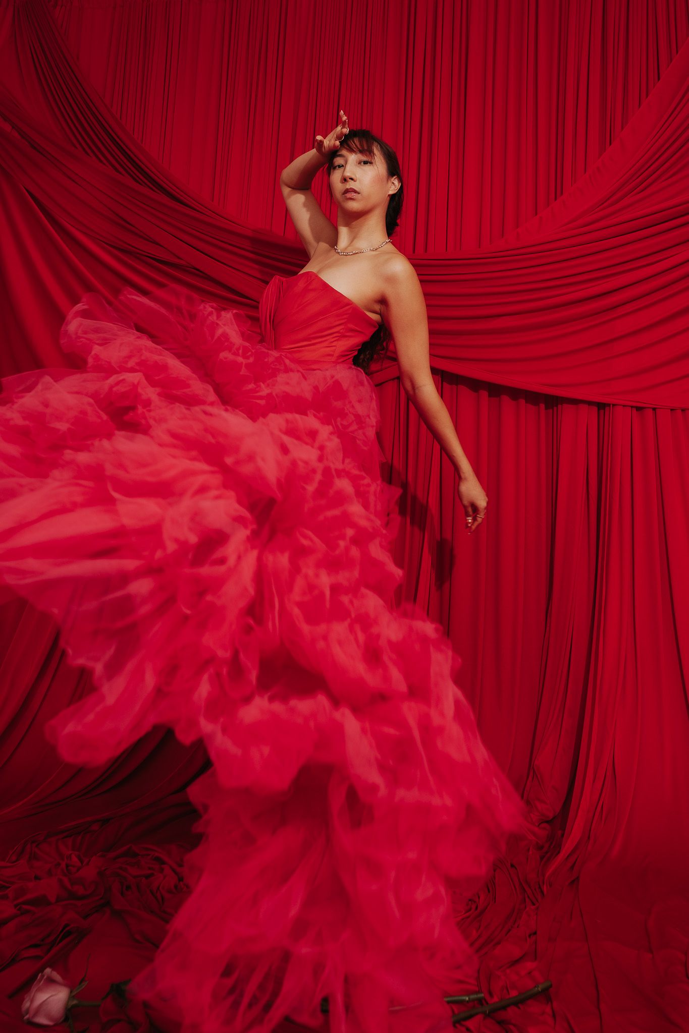 A dramatic fashion photo with a flowing red gown against luxurious red curtain backdrop in theatrical lighting.