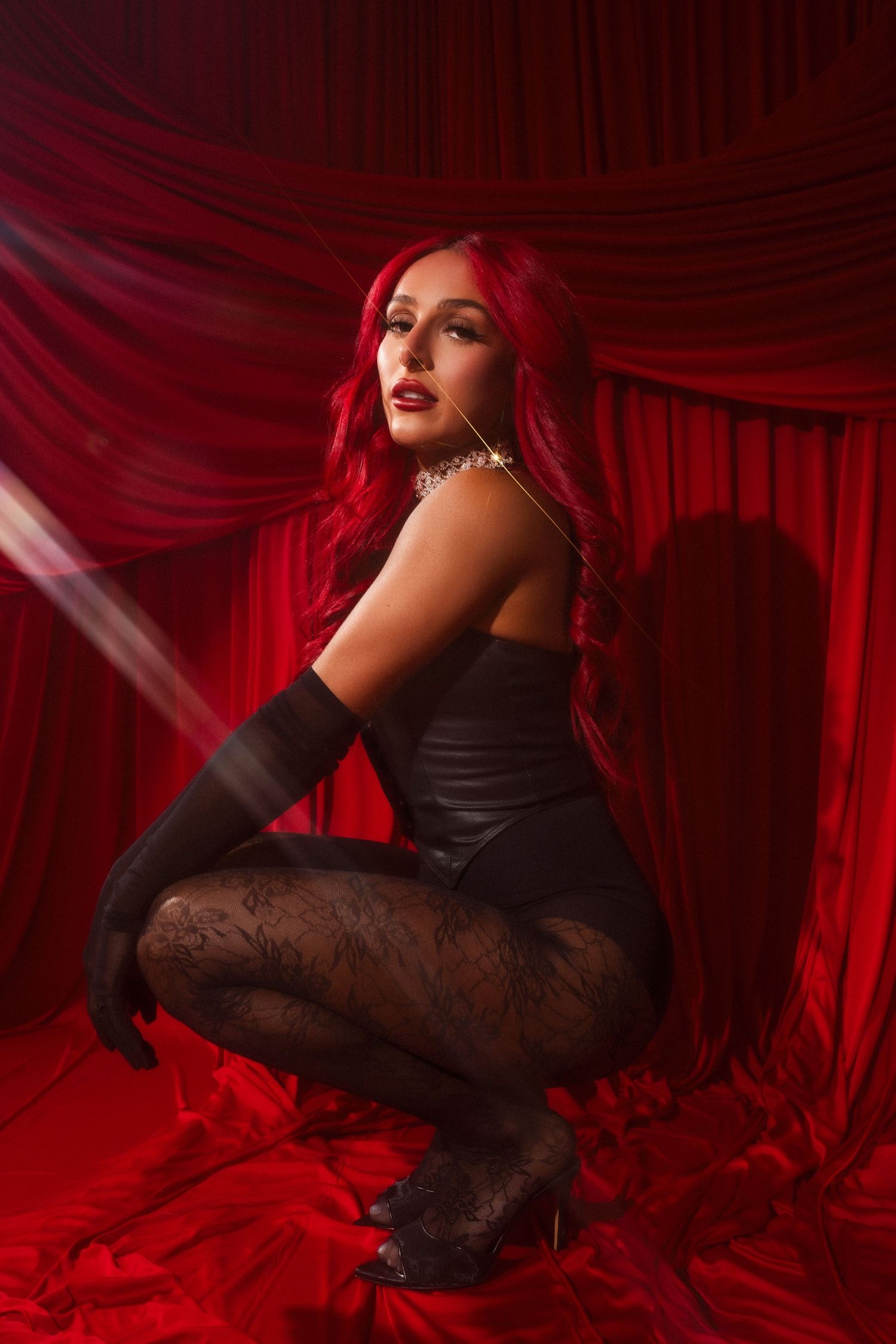 A dark and dramatic boudoir photo with red curtains and soft lighting features a model in black attire and gloves.