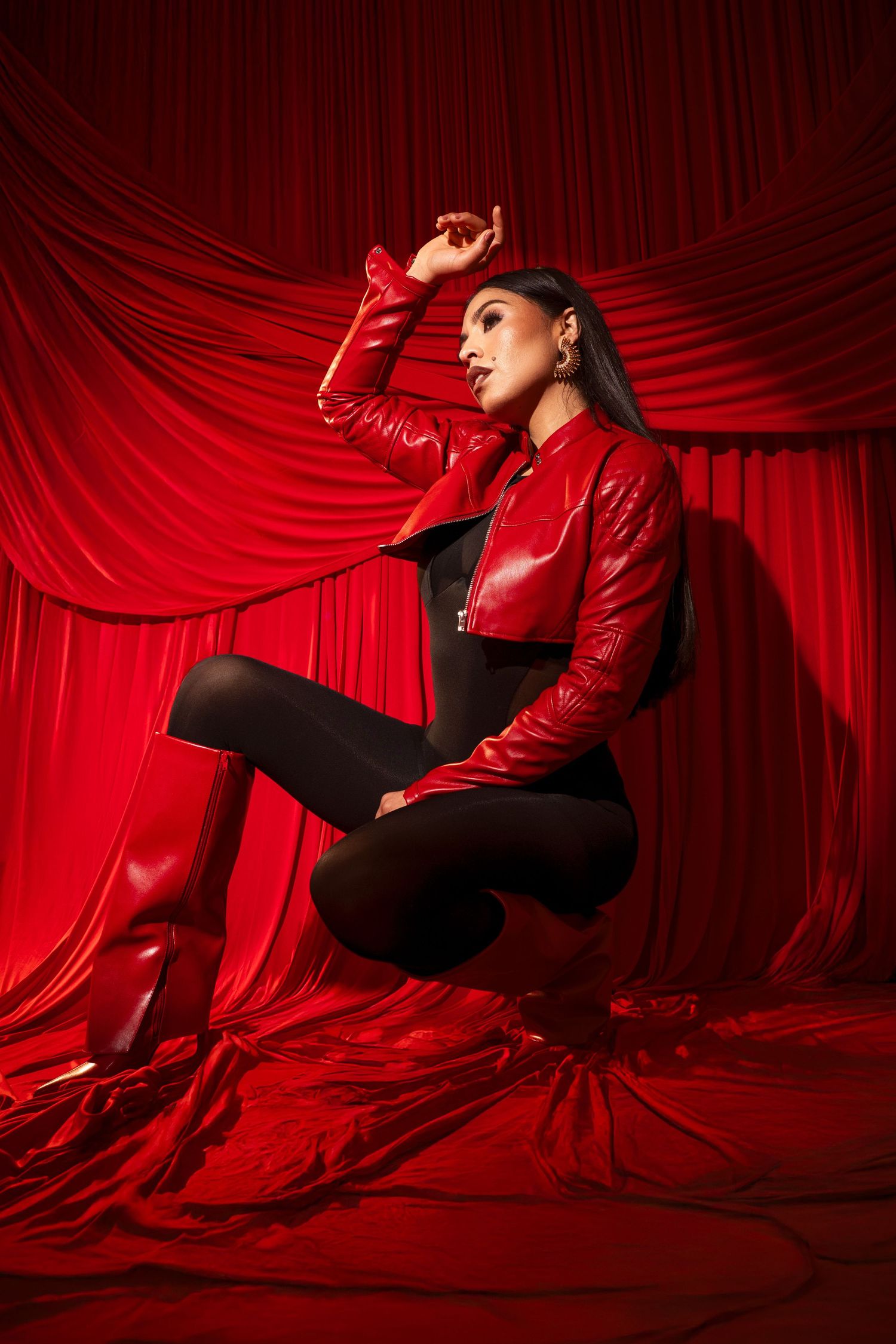 Dramatic fashion portrait against red velvet backdrop with model in shiny red jacket and black pants striking dynamic pose.