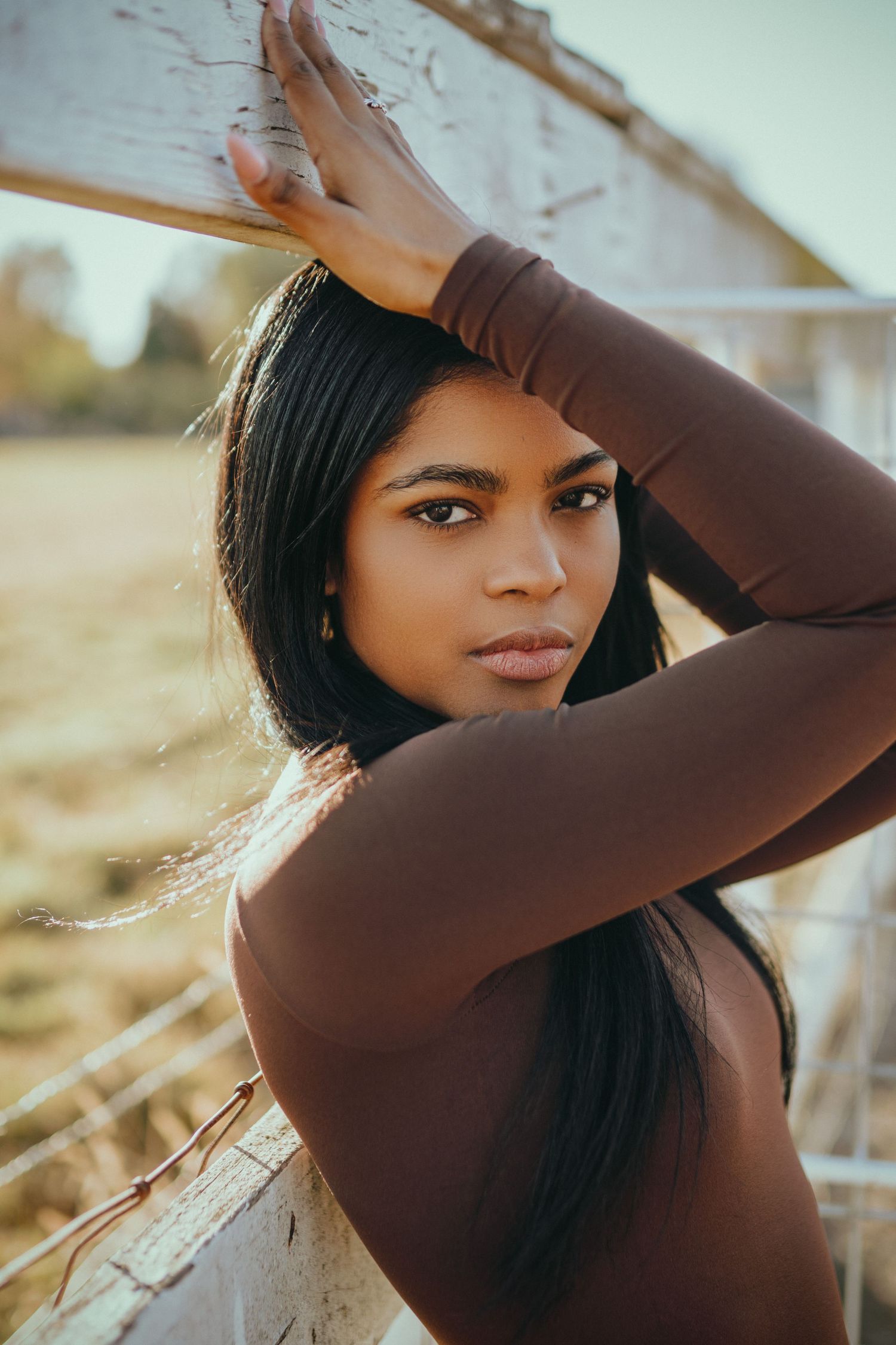 Portrait of a woman in a brown long-sleeved top posing outdoors in golden sunlight against a rustic background.