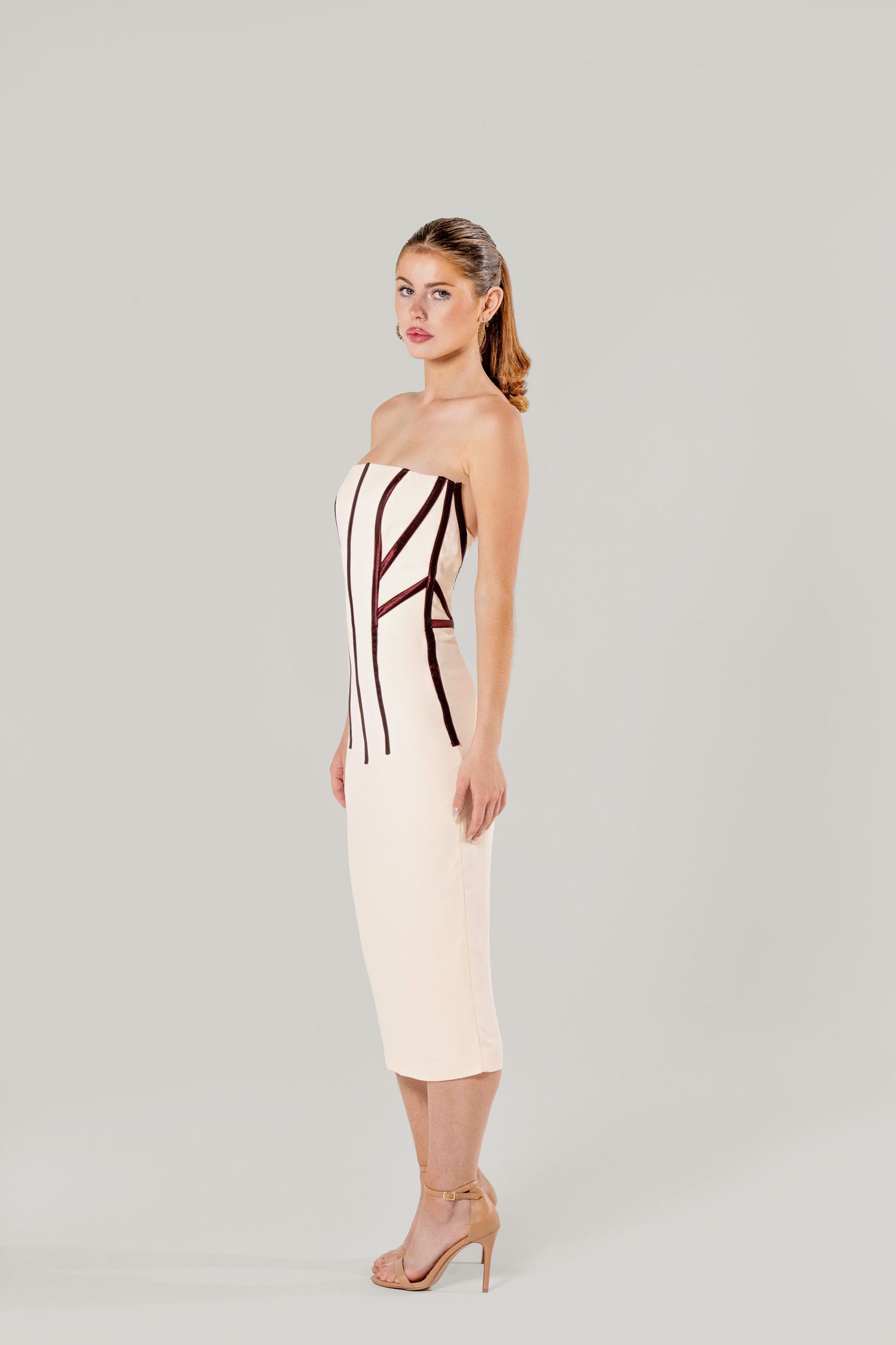 Elegant strapless midi dress in pale pink with black vertical stripes, paired with nude heels in studio setting.