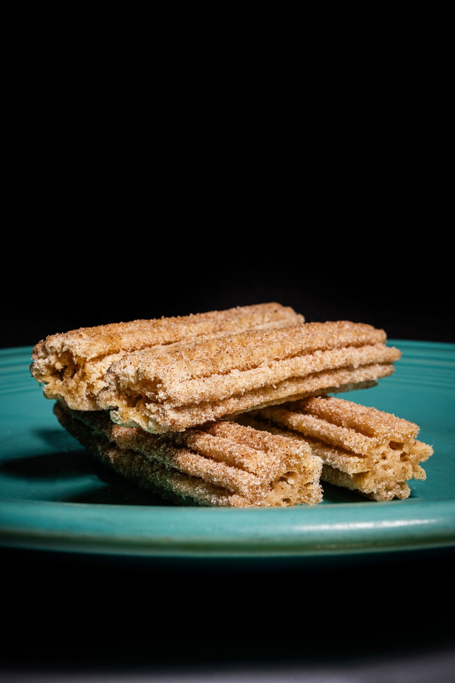 Crispy churros dusted with cinnamon sugar stacked on a turquoise blue plate against a dark background.