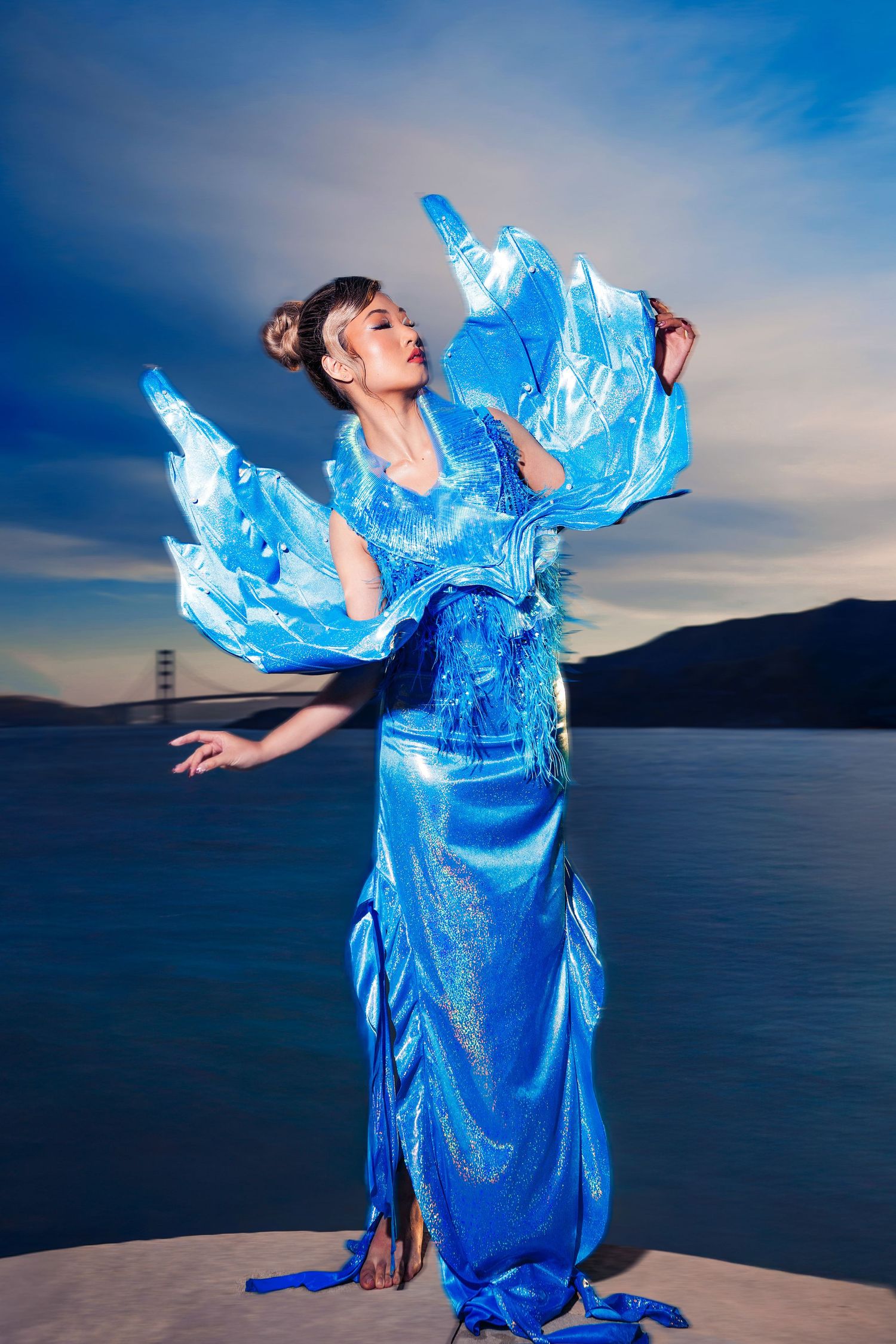 A dancer in flowing bright blue costume with billowing fabric performs against a sunset mountain lake backdrop.