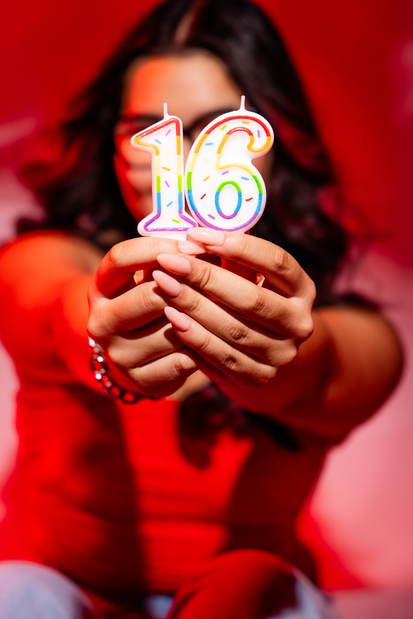 Person holding illuminated rainbow number 16 birthday candle against red background in celebration photo.