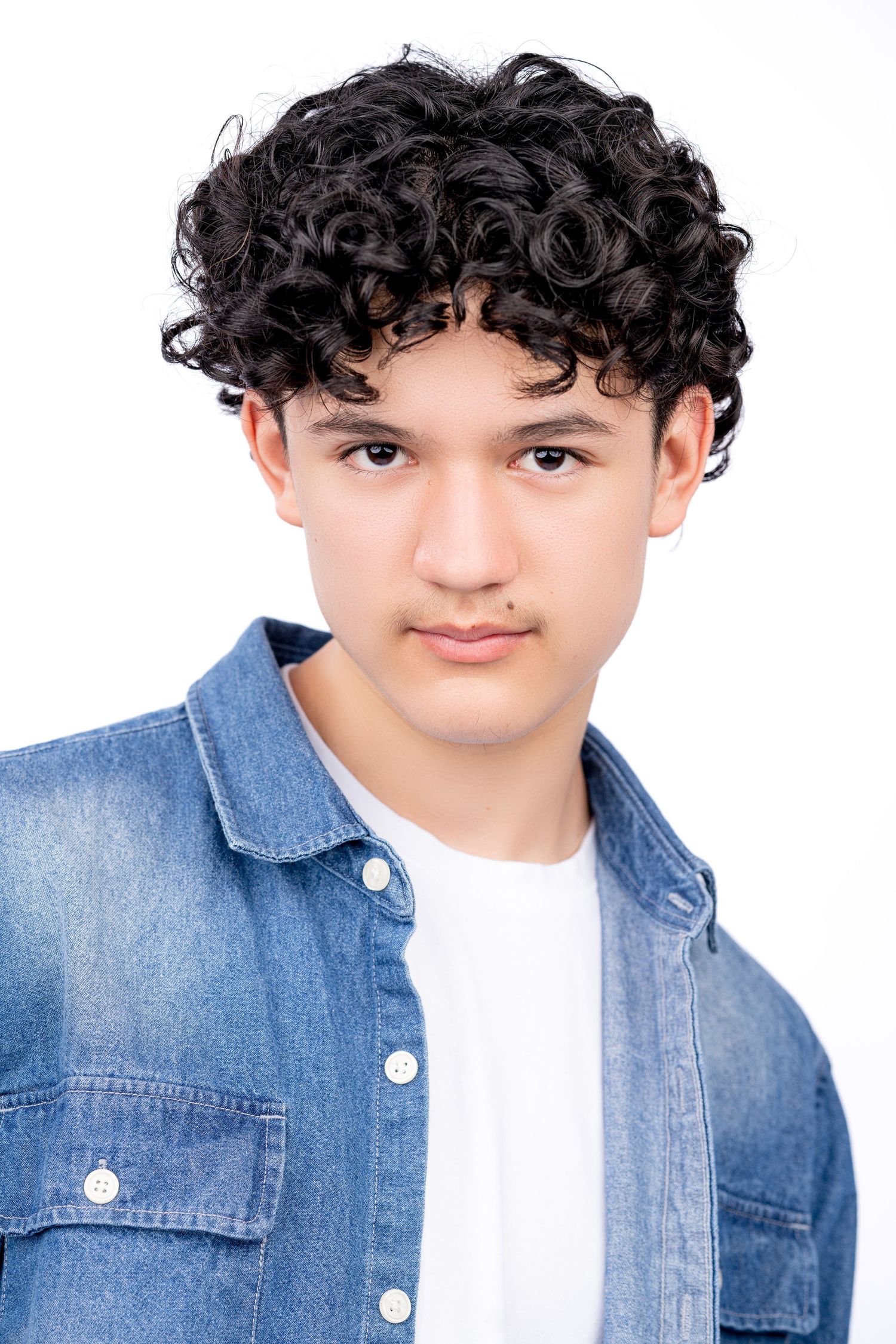 Portrait of a young teenager with dark curly hair wearing a denim button-up shirt over a white t-shirt.