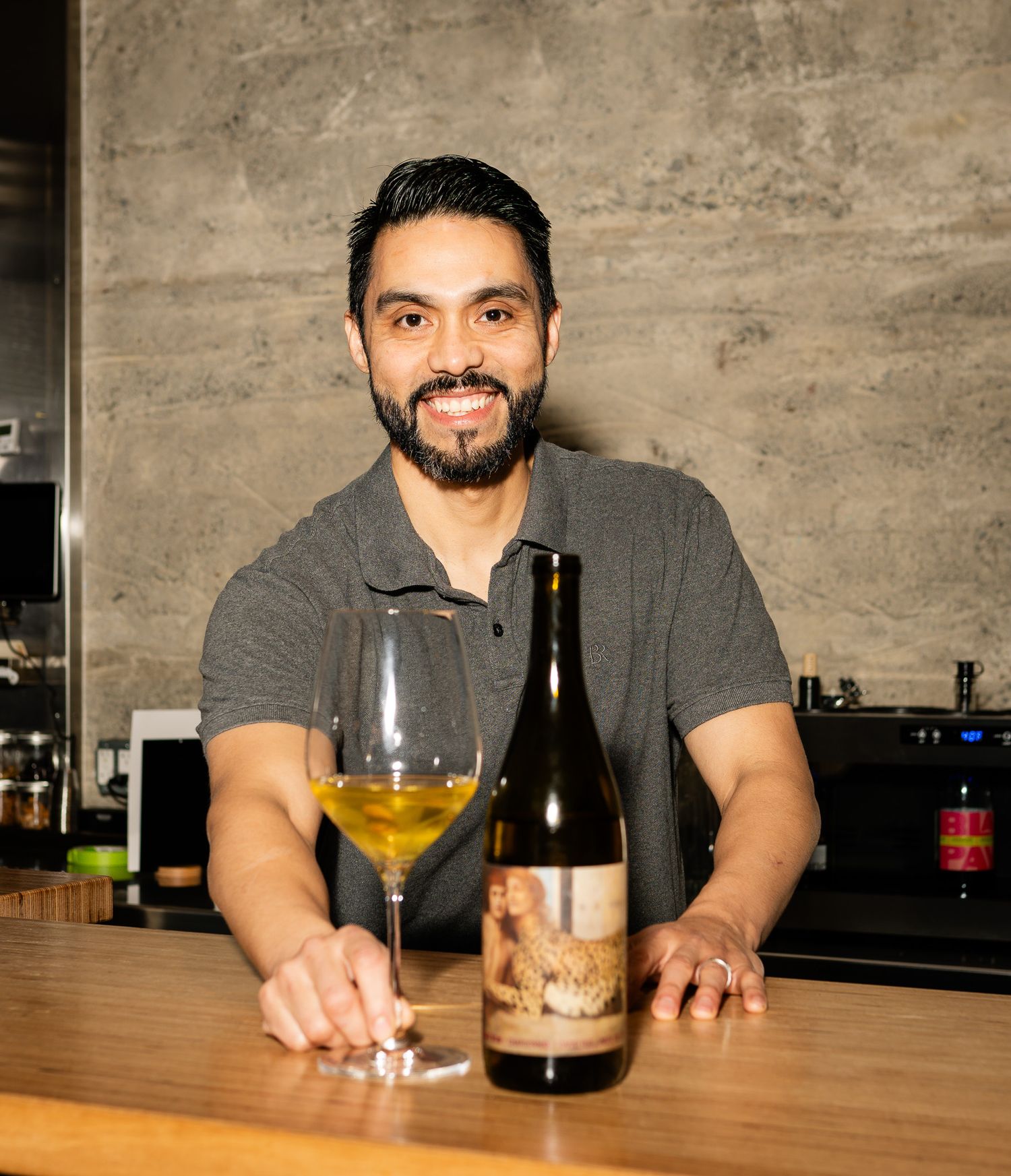 Someone stands behind a bar with a glass of white wine and bottle, wearing a grey polo shirt and smiling warmly.