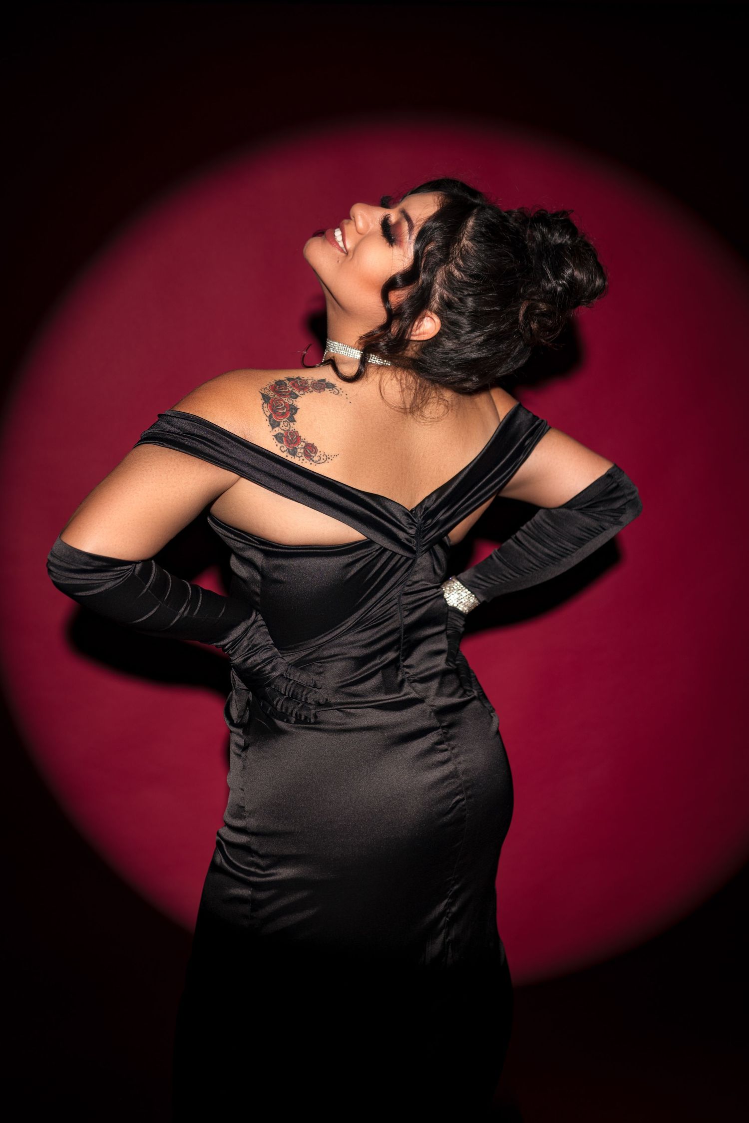 Dramatic portrait of a figure in a black off-shoulder evening gown against a deep red circular background.