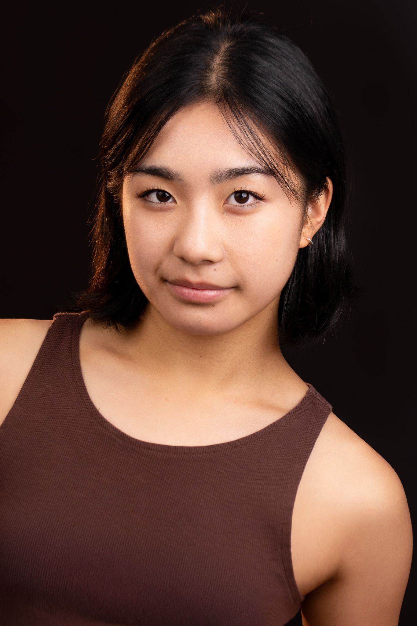 Young woman with short dark hair poses against dark background wearing a brown sleeveless top.
