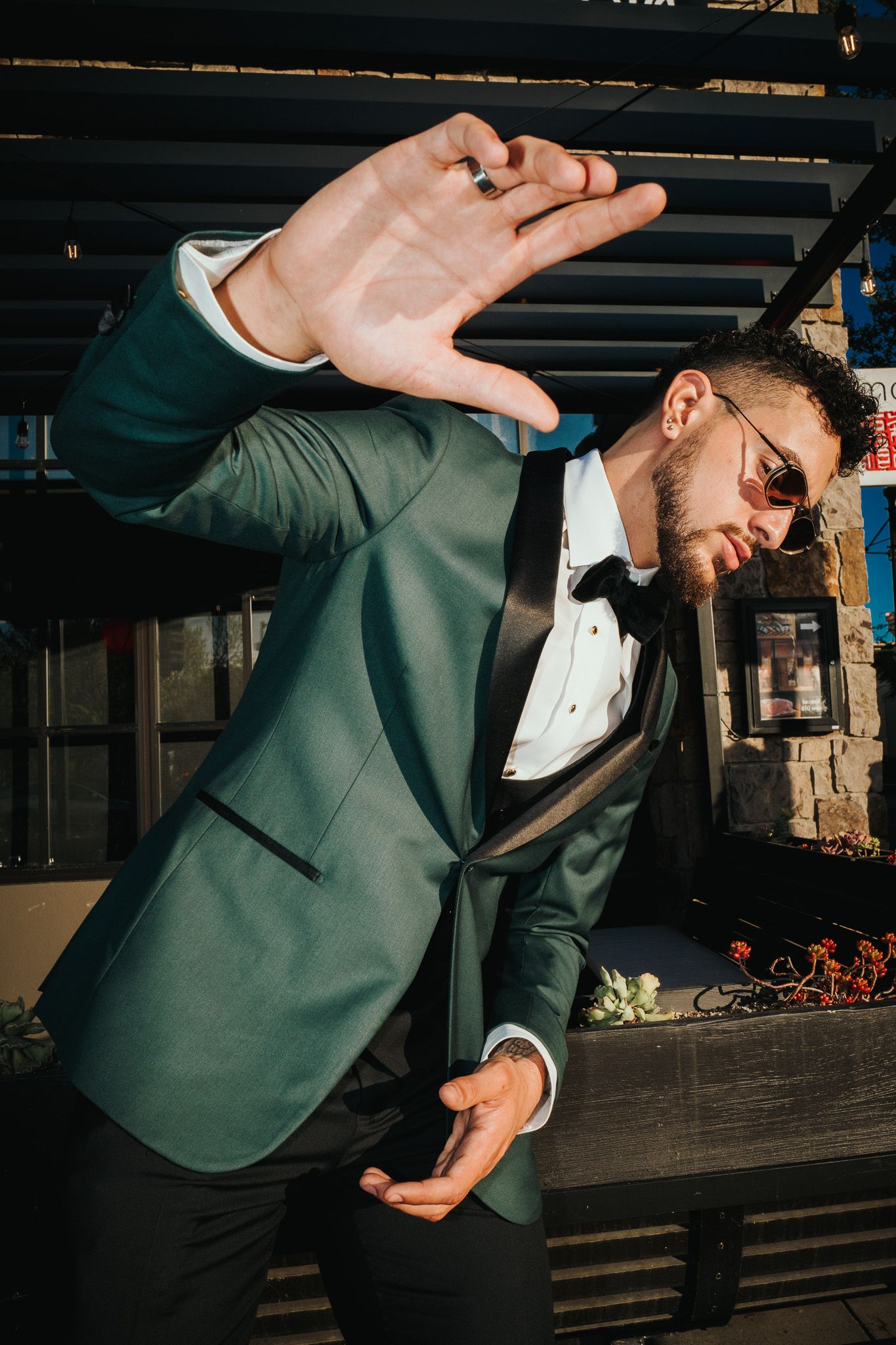 A person in a forest green tuxedo jacket and sunglasses strikes a dynamic dance pose against an industrial backdrop.