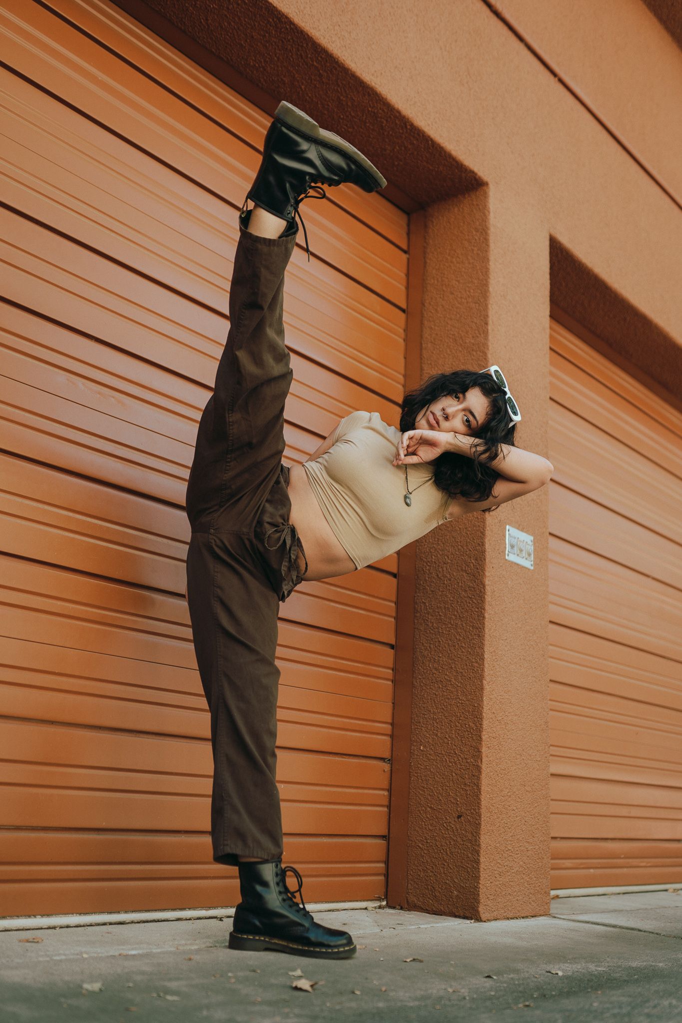 A person in casual attire performs a high split kick against an orange wall with rolling doors.