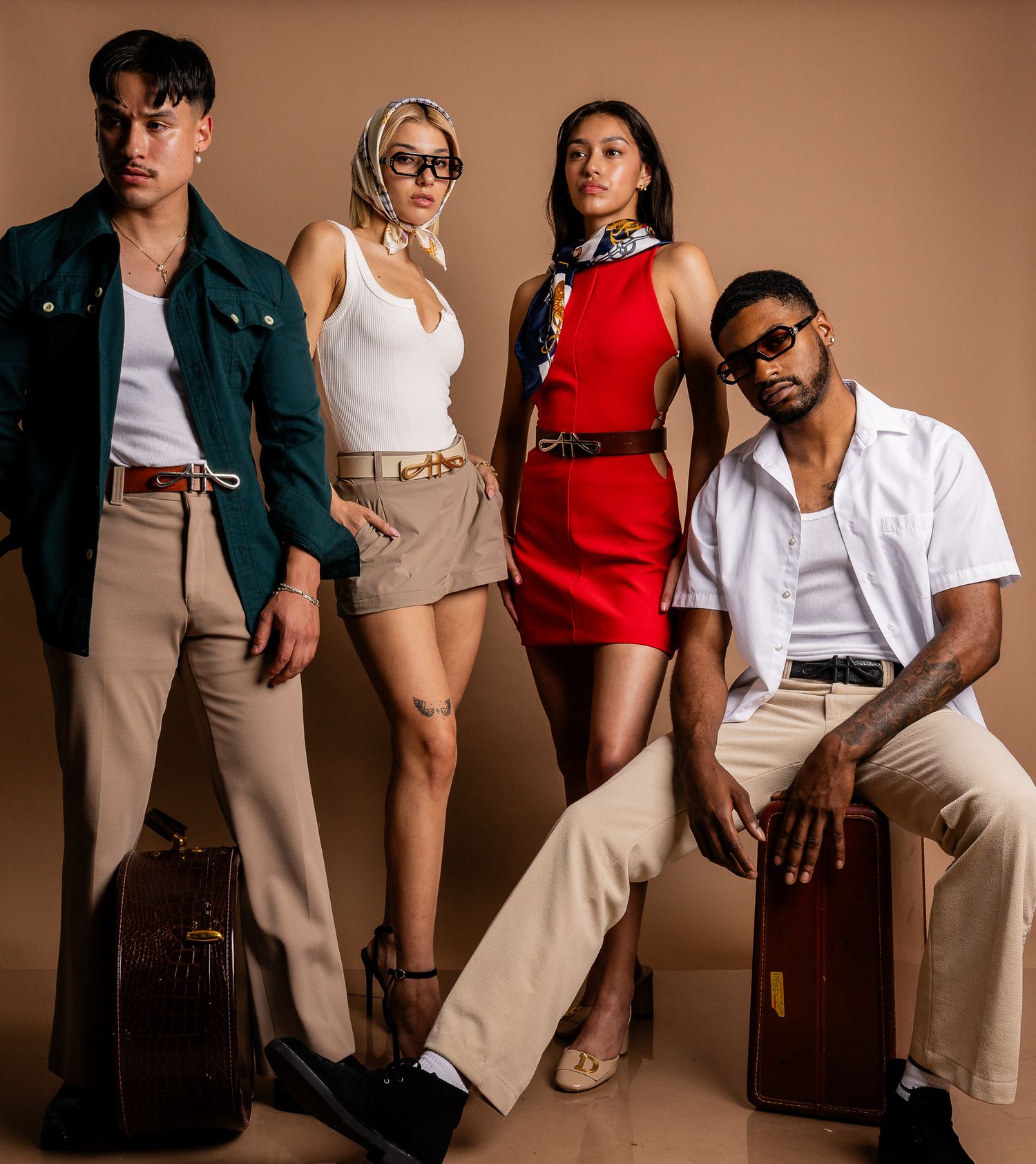 Four models in stylish casual and formal wear pose on brown suitcases against a neutral backdrop in a fashion photo shoot.