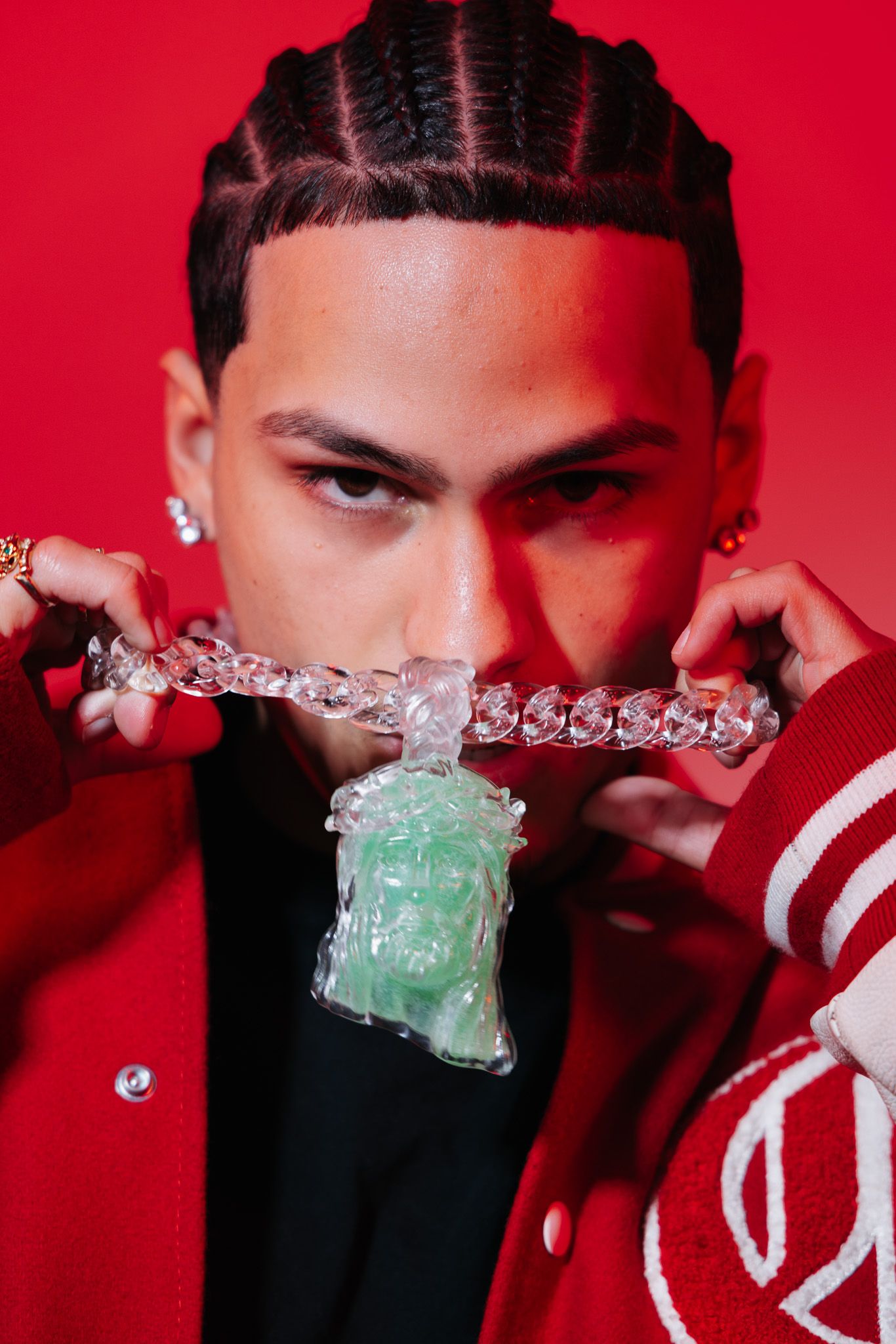 A person wearing braids and a red jacket displays a large diamond chain necklace with a jade pendant against a red background.