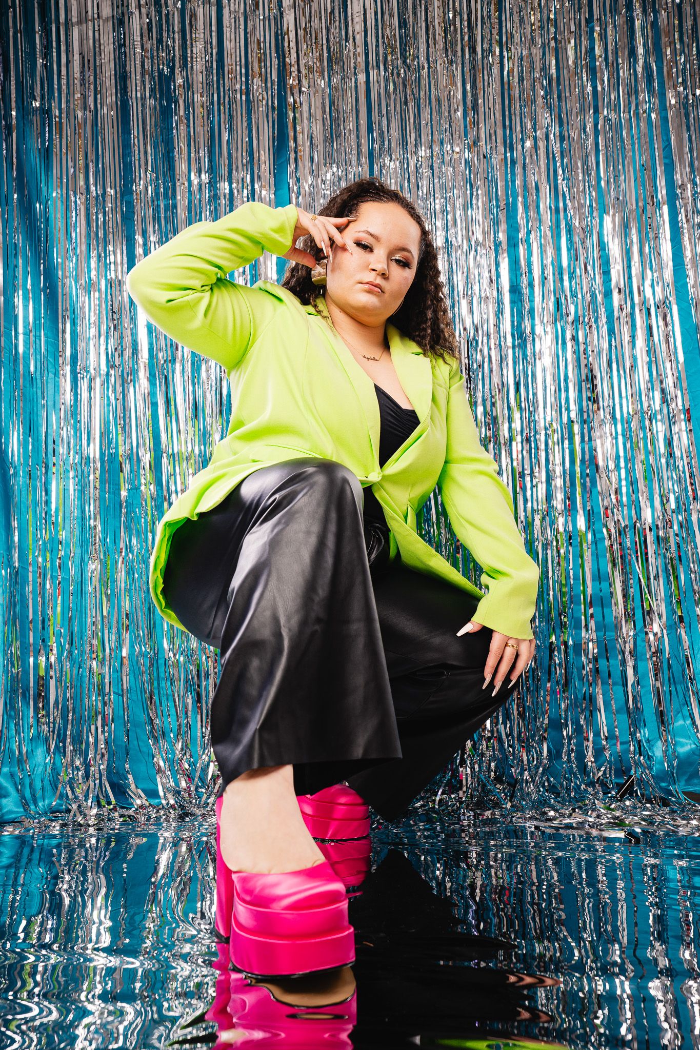 A model poses in a neon yellow jacket and pink boots against a metallic blue tinsel backdrop.