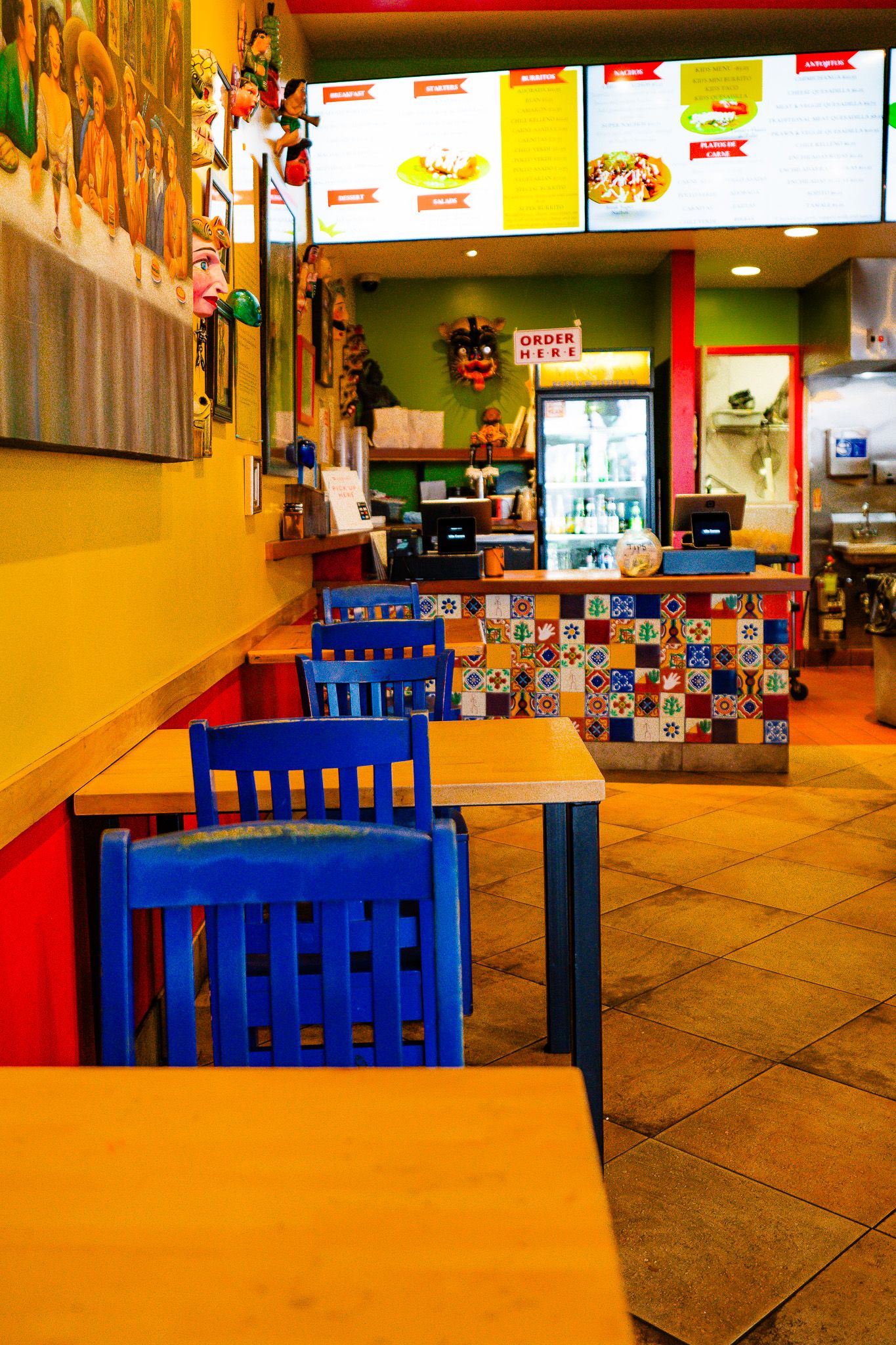 Colorful Mexican restaurant interior with bright blue chairs, yellow walls and menu boards displaying food items.