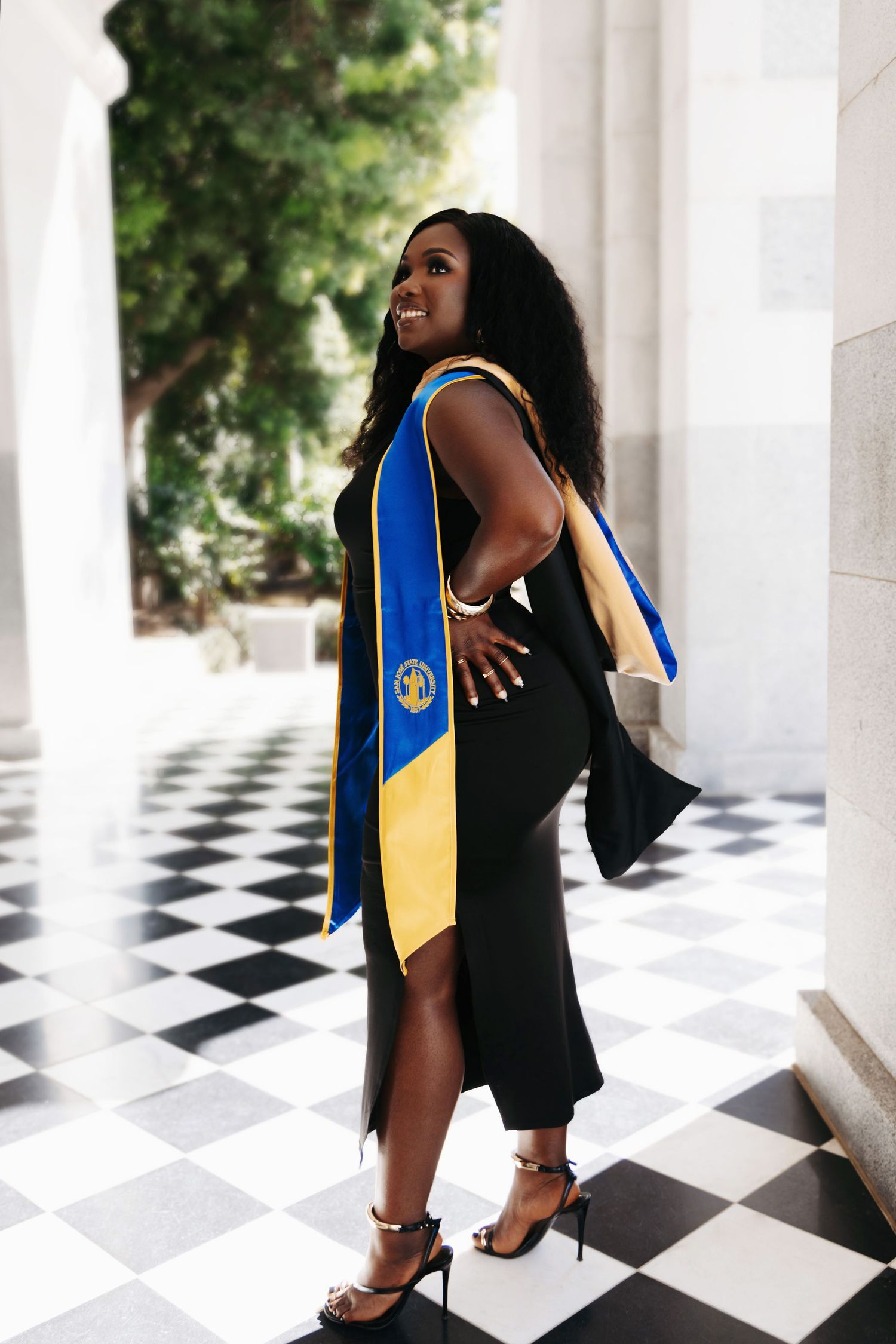 Graduate poses in a black dress with blue and gold academic stole on checkered floor during graduation photoshoot.