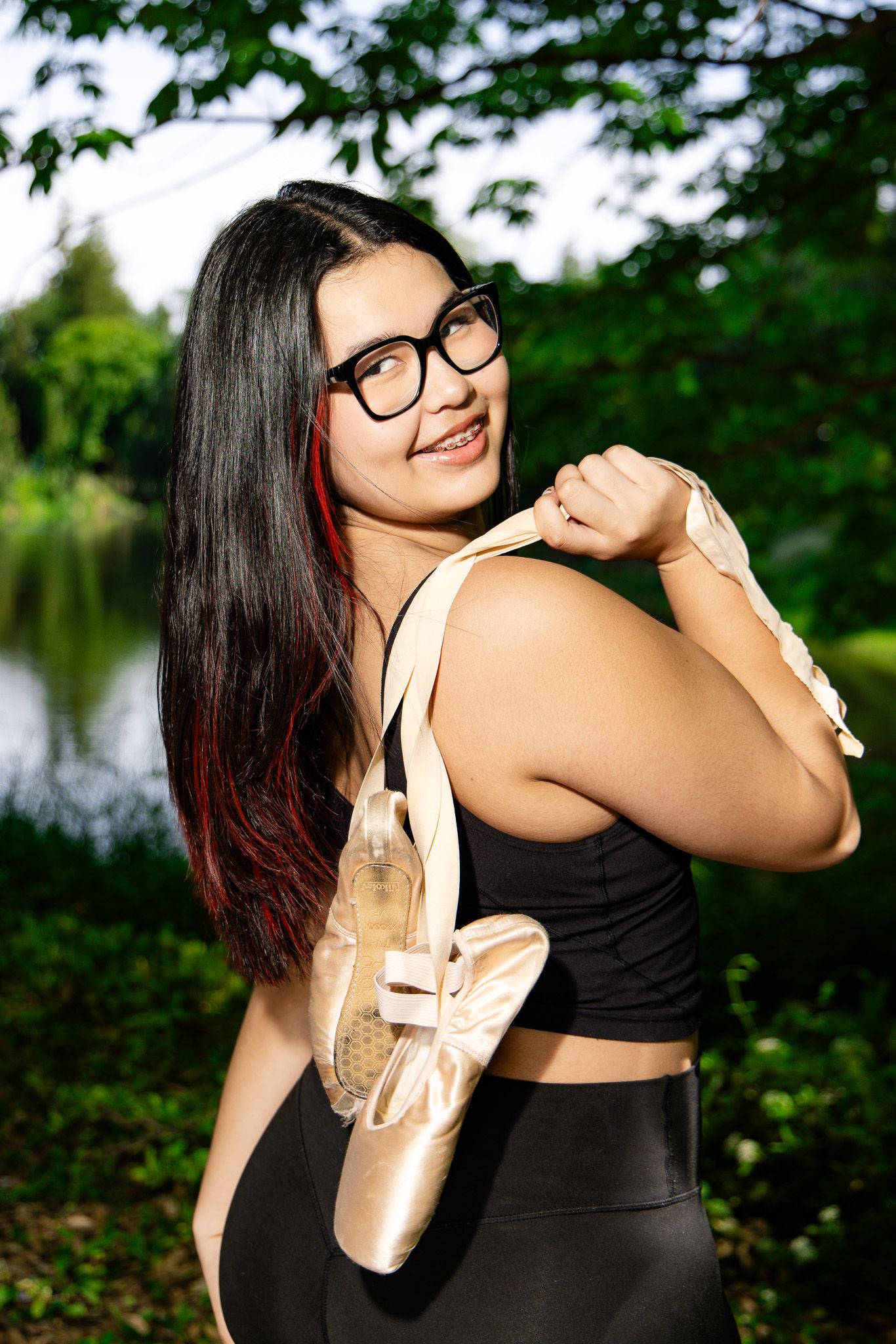 A dancer in black athletic wear holds ballet pointe shoes outdoors by a lake with greenery in the background.