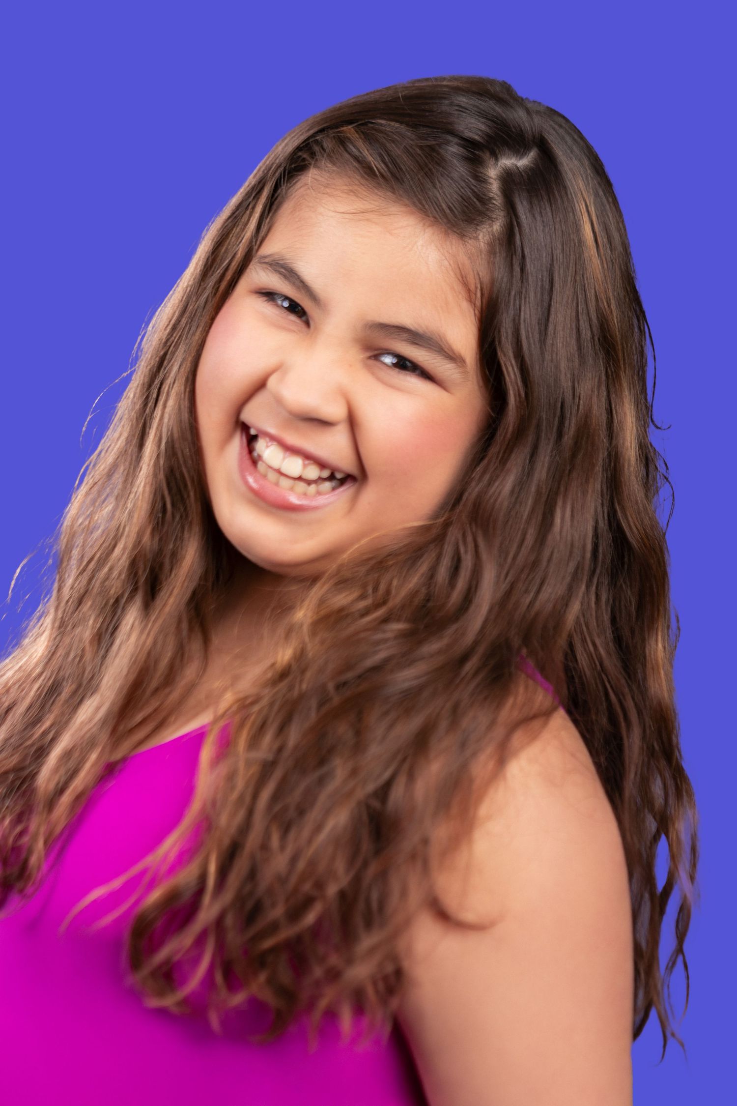 Joyful portrait of someone with long brown wavy hair wearing a bright pink top against a vivid blue background.