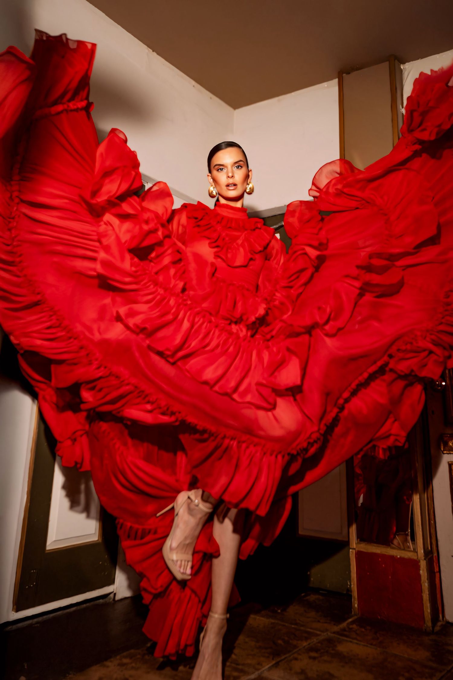 A dramatic fashion photograph featuring a flowing red ruffled dress in motion against a white wall.