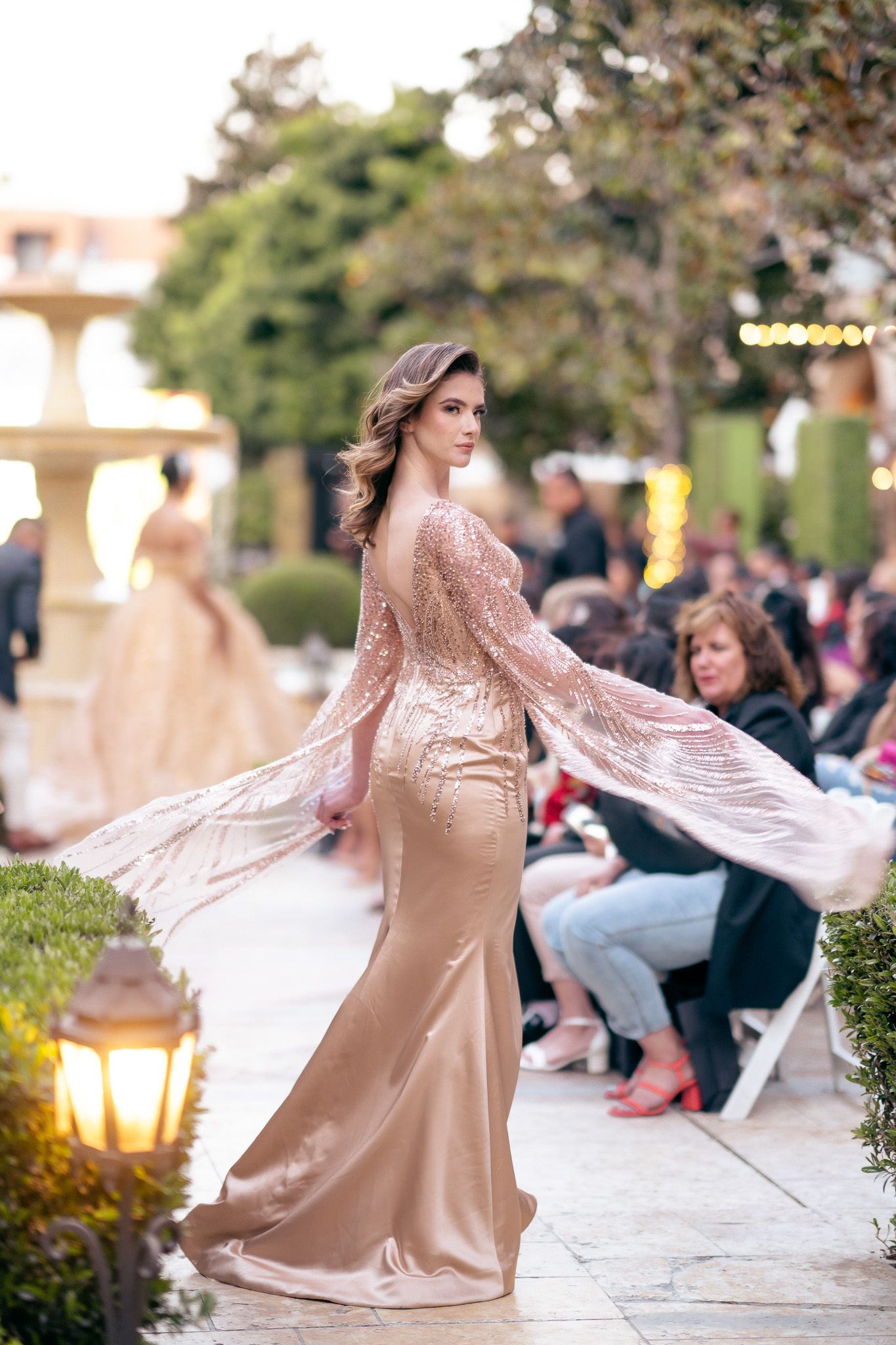 Model walks runway in flowing champagne-colored evening gown with lace overlay at outdoor fashion show.