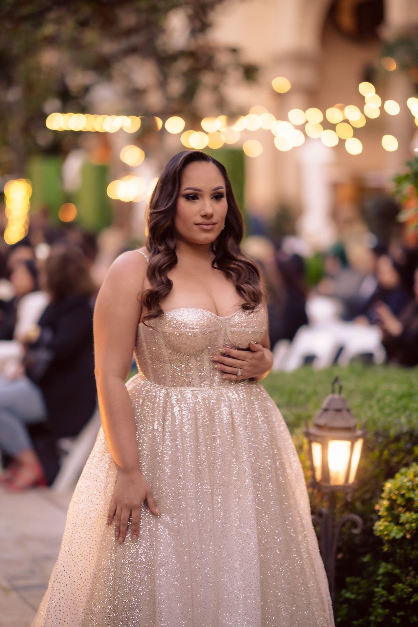 A woman in a sparkly champagne-colored ball gown stands at an outdoor evening event with twinkling lights in the background.