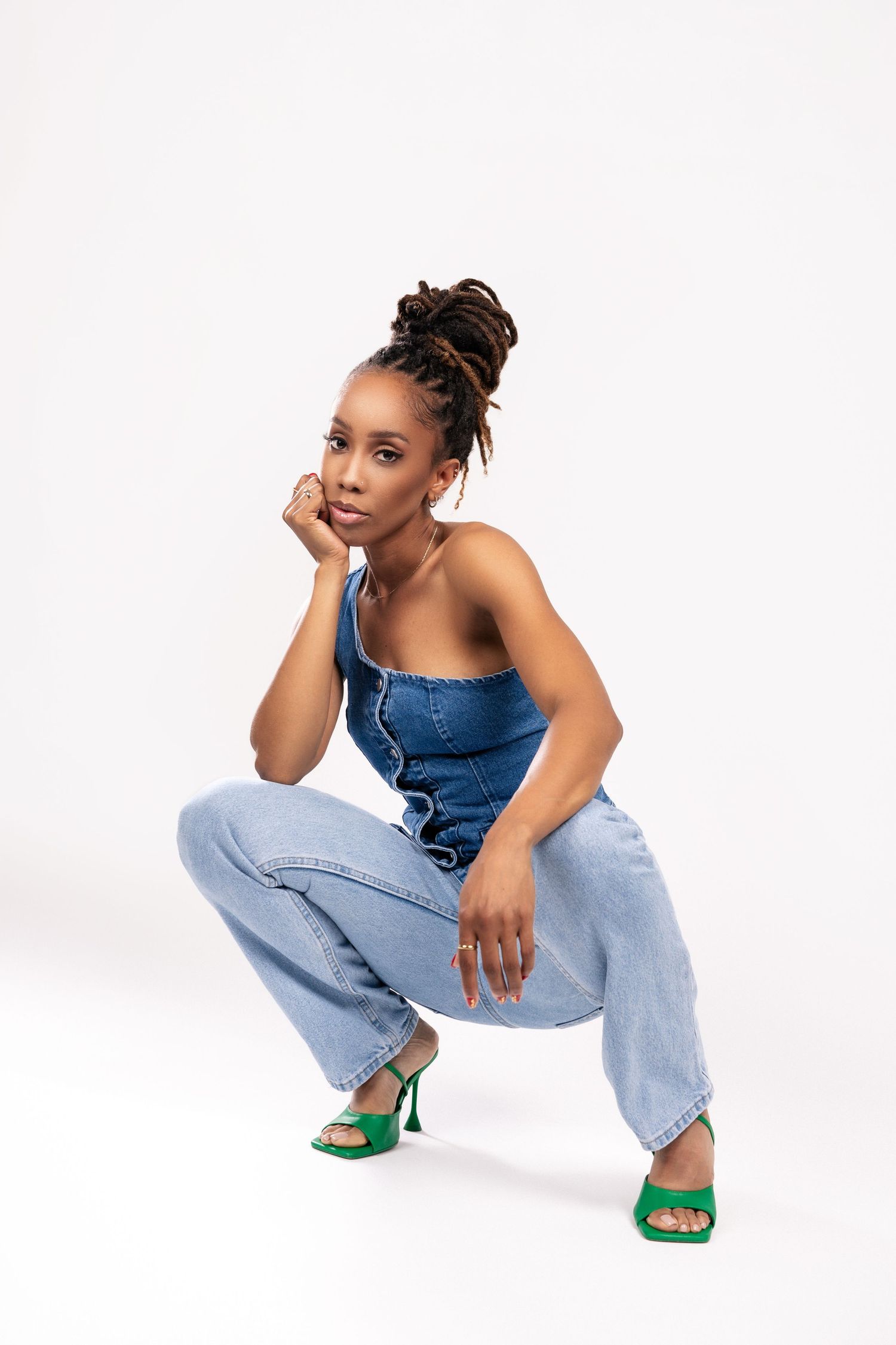 Person in casual denim outfit with stacked bun hairstyle crouches in studio wearing blue top and jeans with green heels.