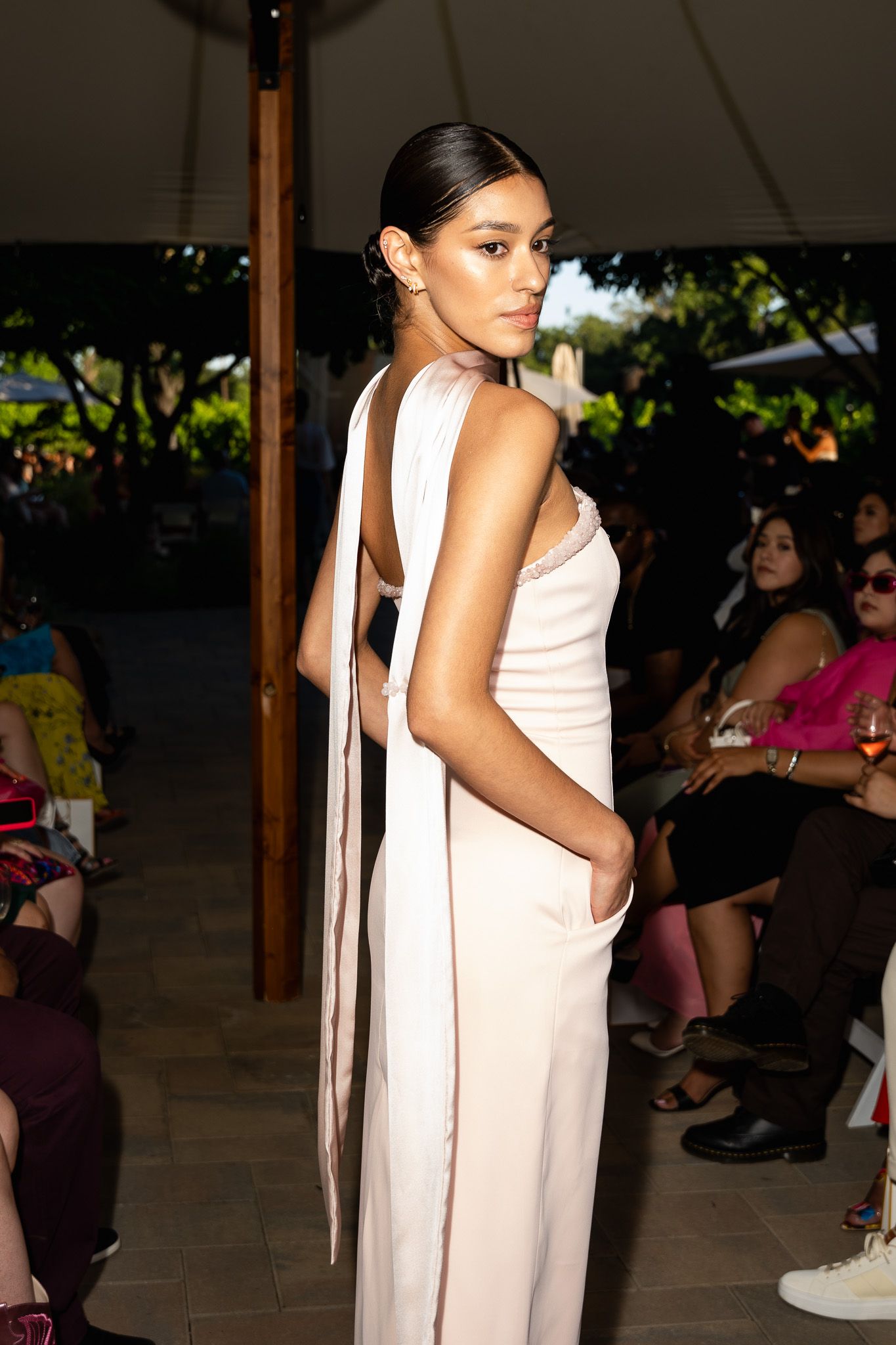 Model wearing an elegant pale pink jumpsuit with trailing back ribbons on a fashion runway with audience in background.