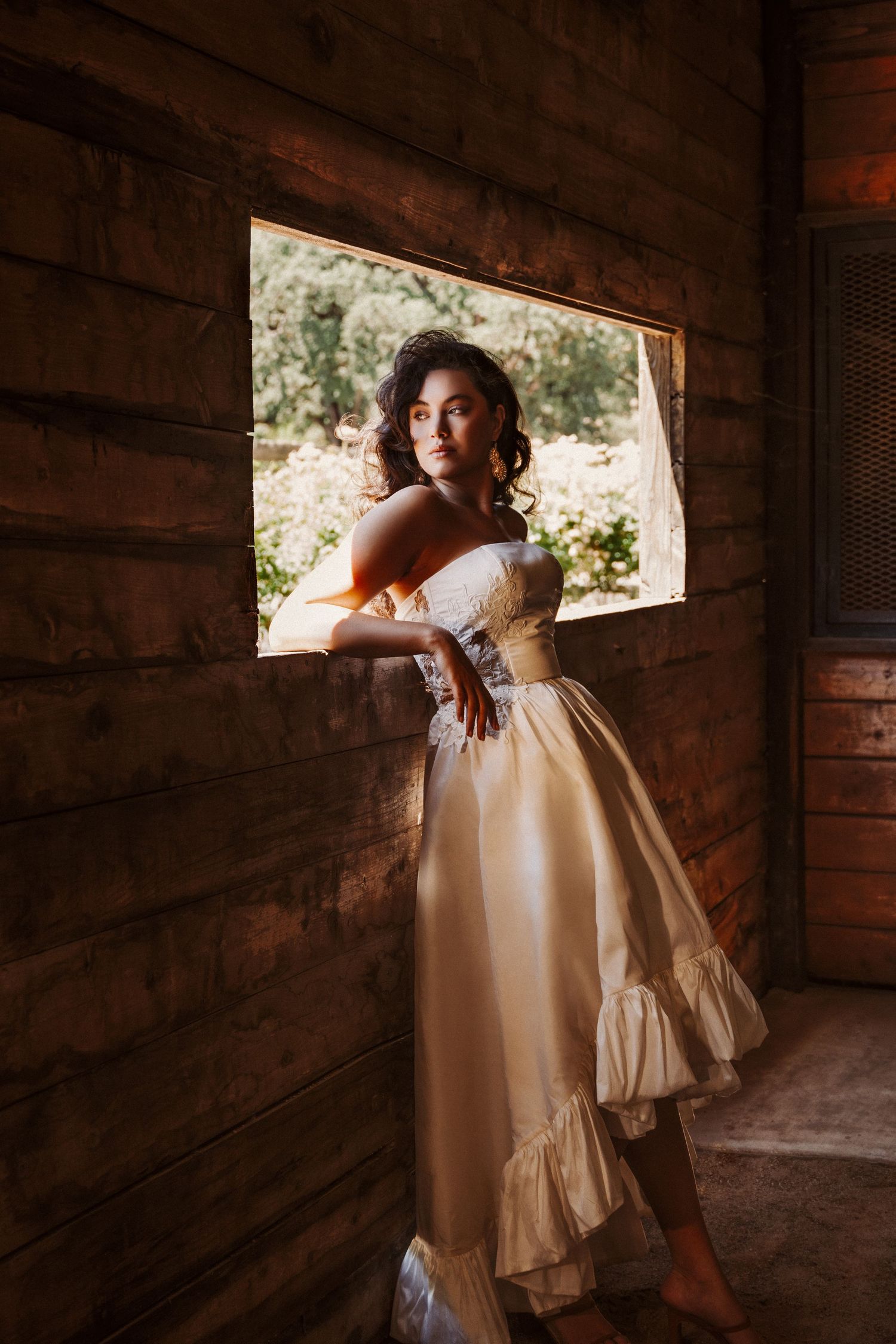 A person in a vintage cream-colored dress poses by a rustic cabin window in warm natural lighting.