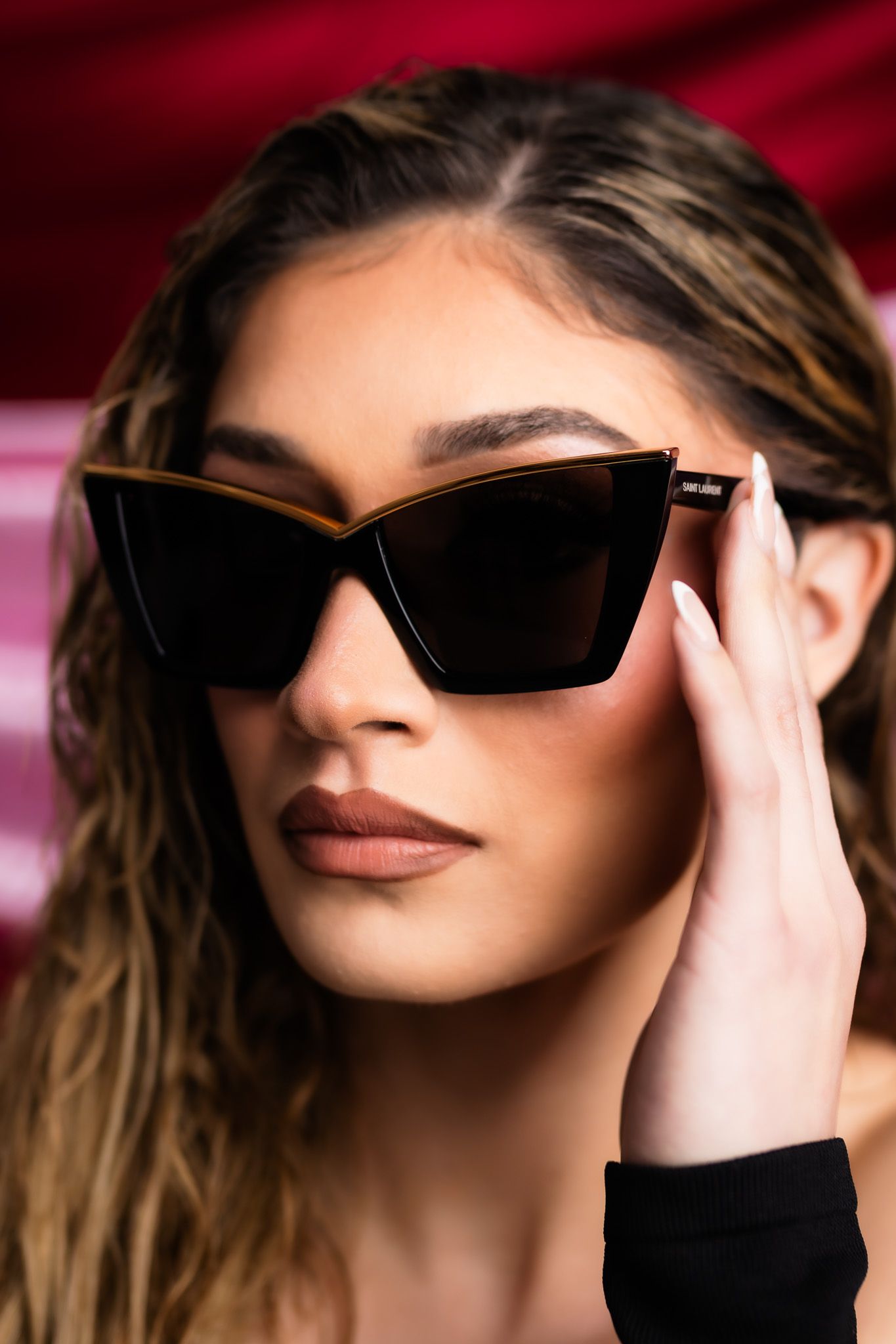 Stylish fashion model in black cat-eye sunglasses poses against red backdrop with hand touching frame.