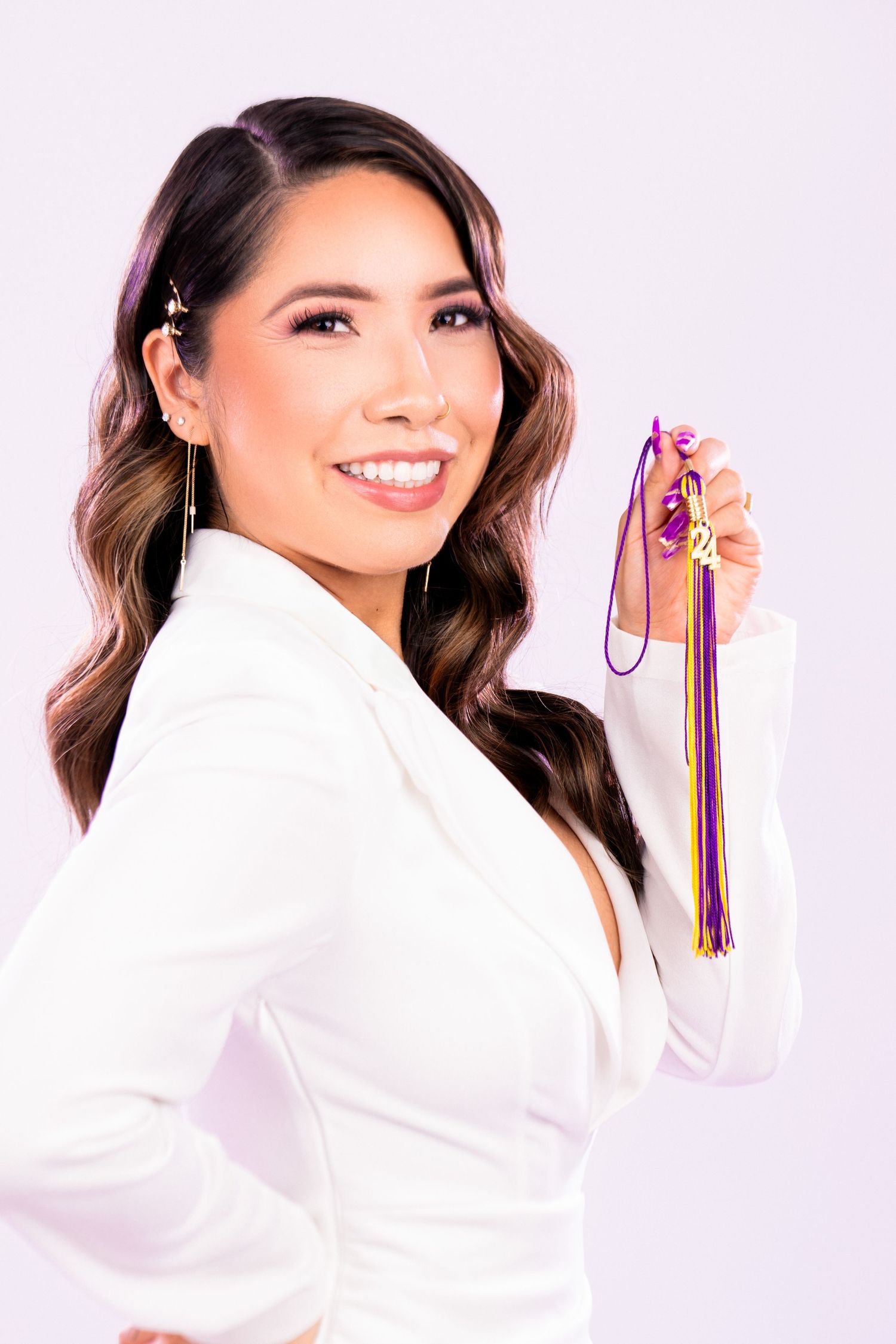 Woman in white blazer holding colorful ribbons while posing against light pink background with wavy brown hair.