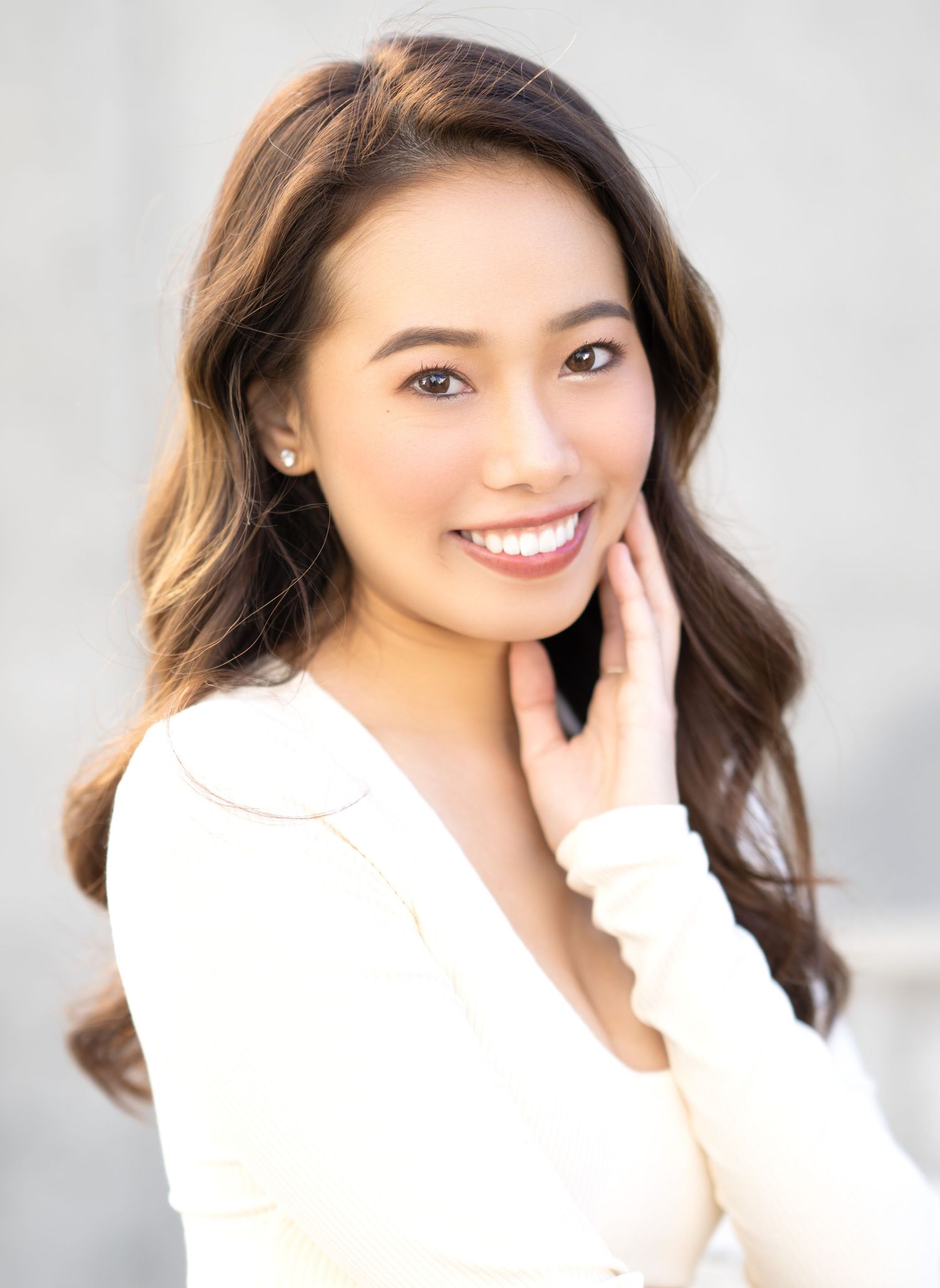 Professional portrait of a person with long wavy brown hair wearing a white sweater smiling warmly against a gray background.