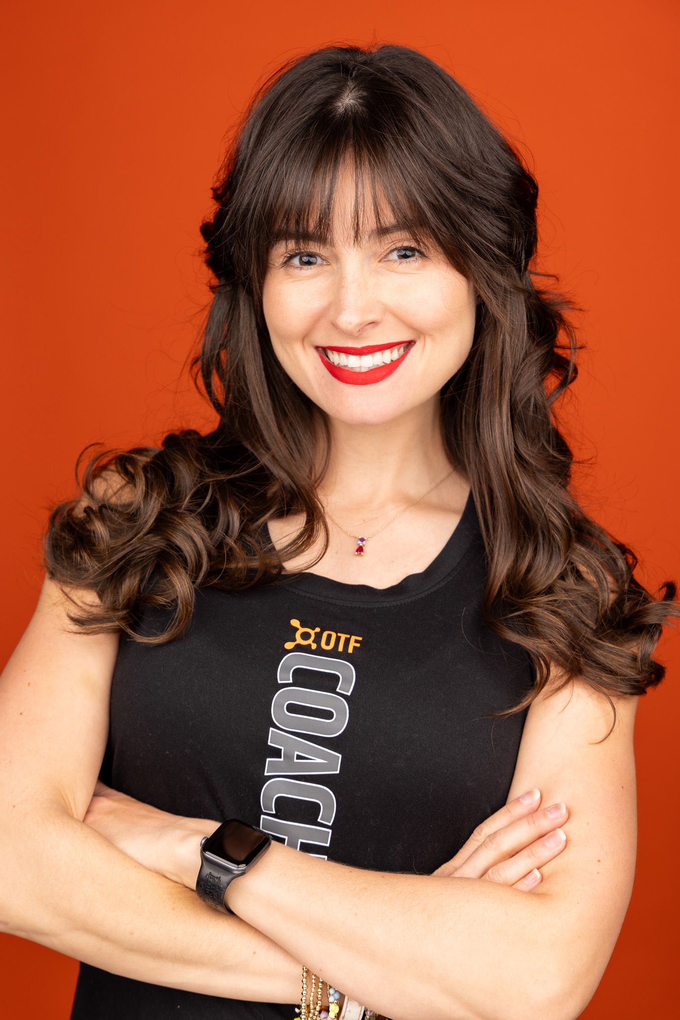 Portrait of a smiling person with long dark wavy hair wearing a black tank top against an orange background.