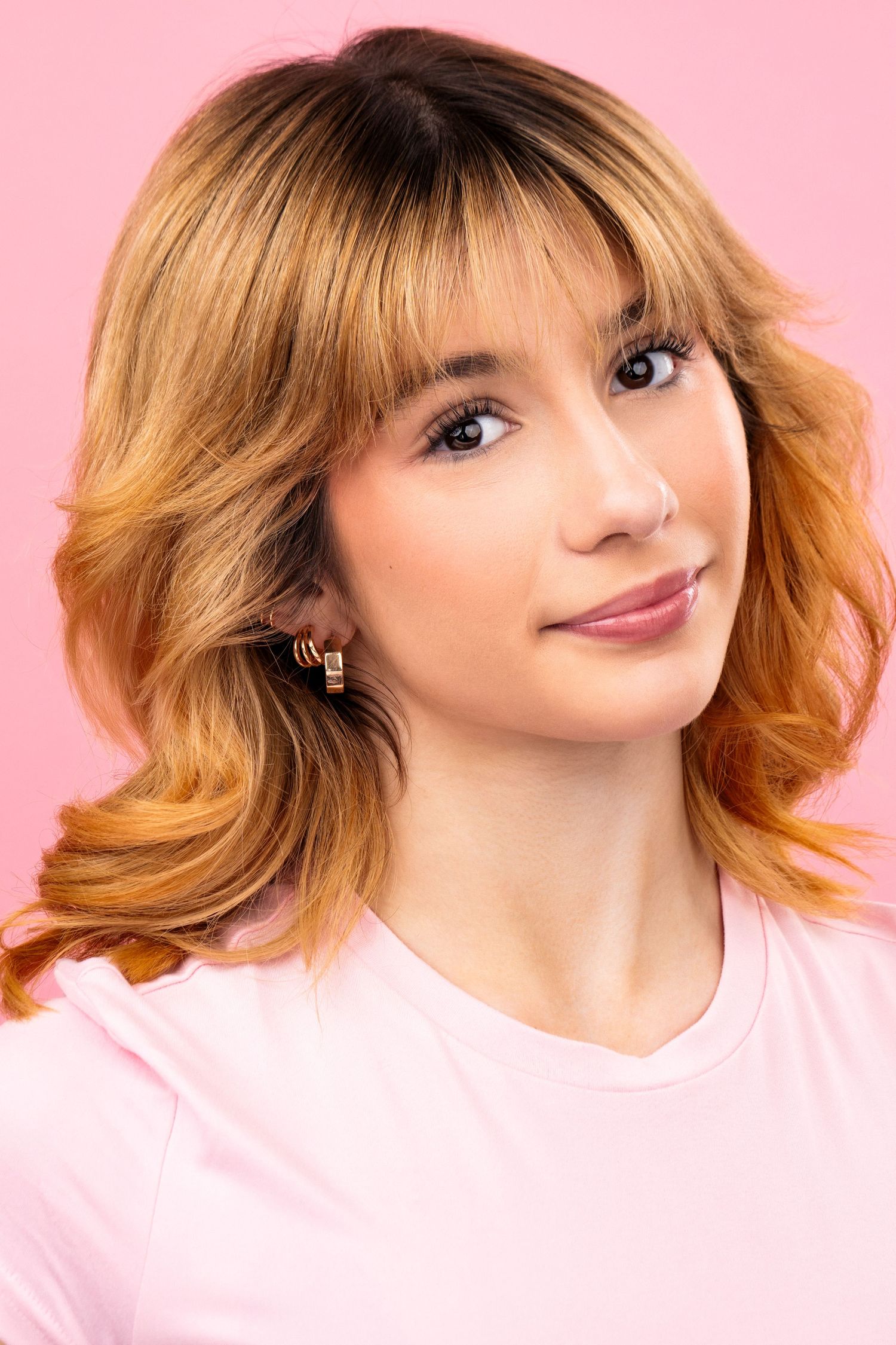 Portrait against pink background showing honey blonde layered hair and soft smile while wearing a pink shirt.