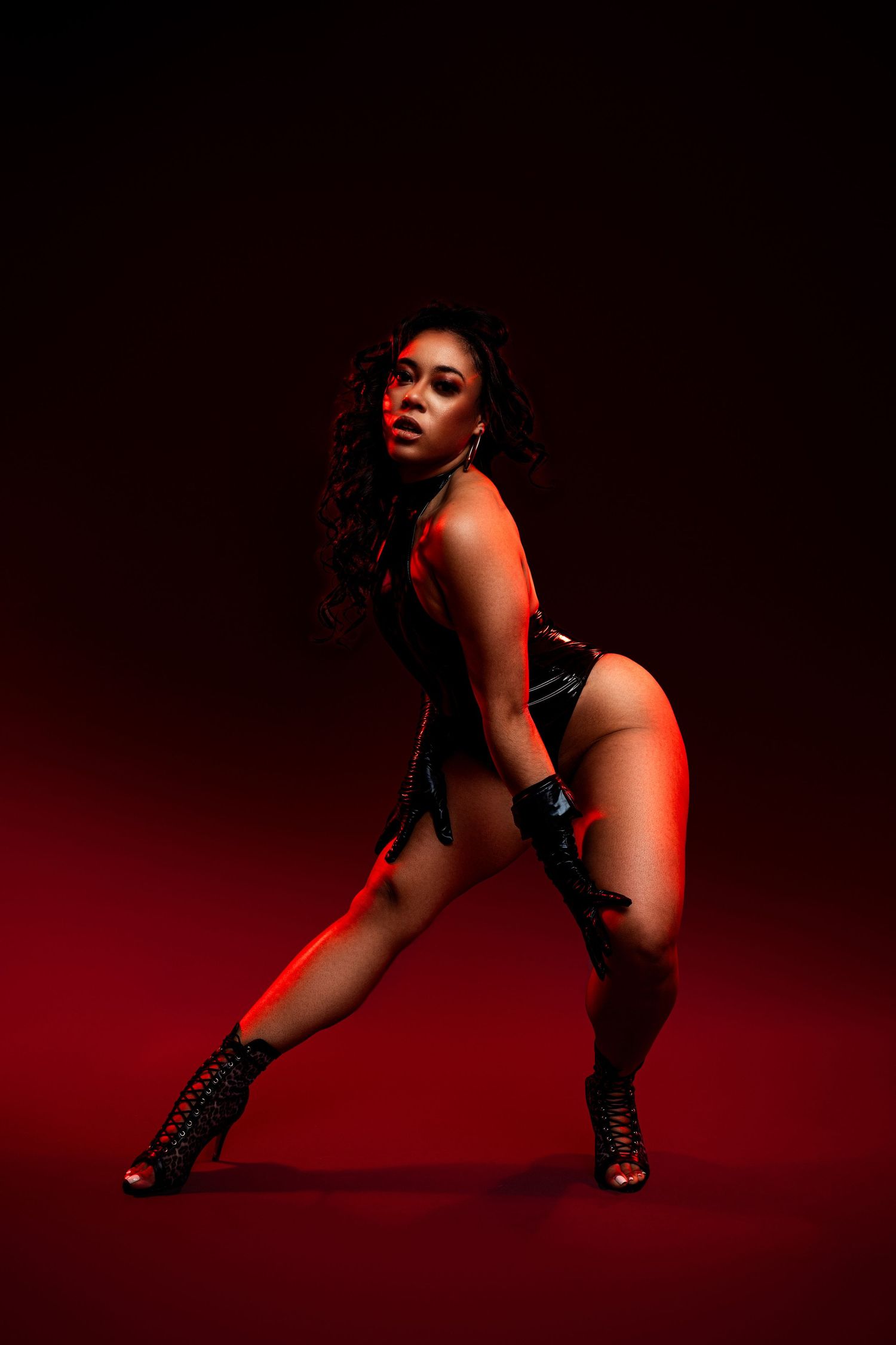 Dramatic portrait of a dancer in black bodysuit and boots striking a powerful pose against a dark red background.