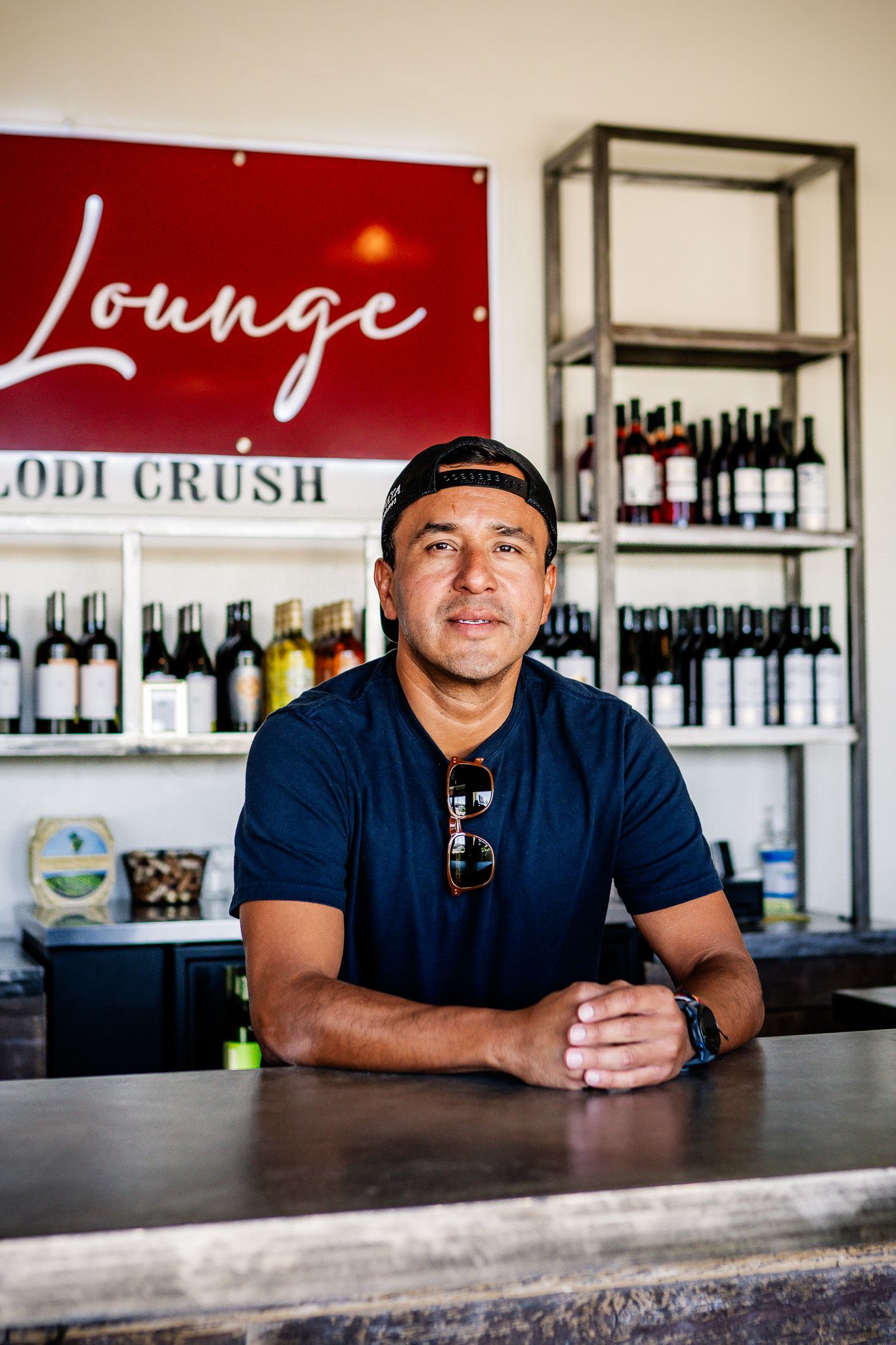 Staff member leans on bar counter at wine lounge with red sign and bottles displayed on shelves in background.
