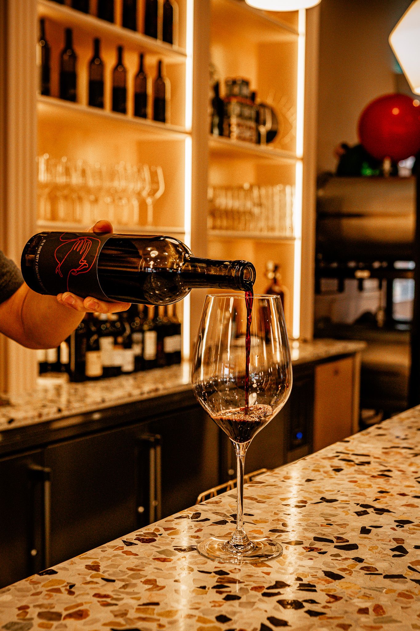 Red wine being poured into a glass at a bar with shelves of bottles and glassware in the background.
