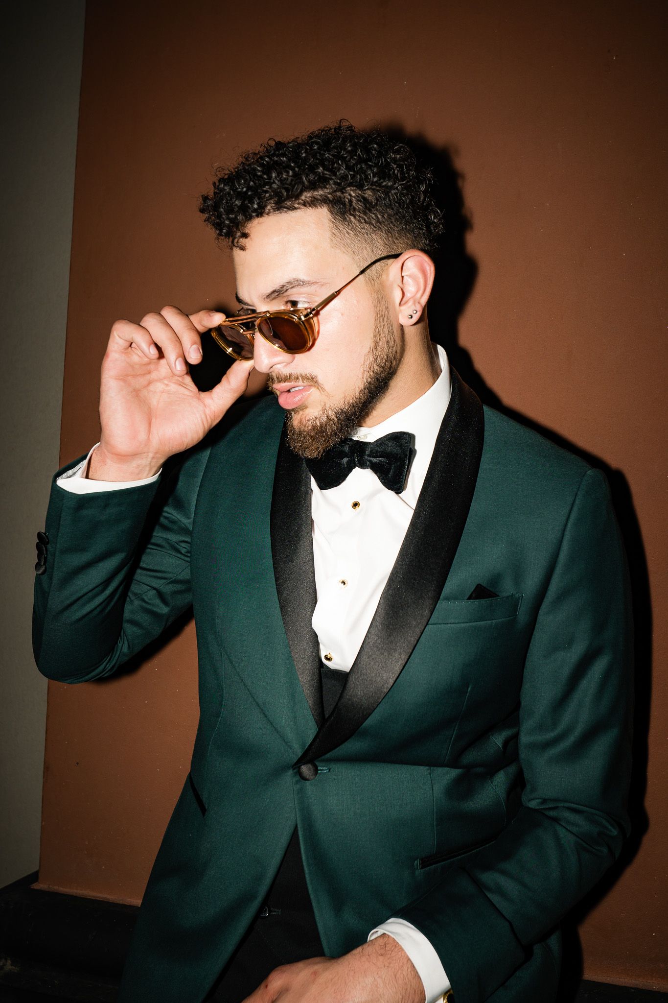 A person in a dark green tuxedo jacket and bow tie adjusts their sunglasses against a brown background.