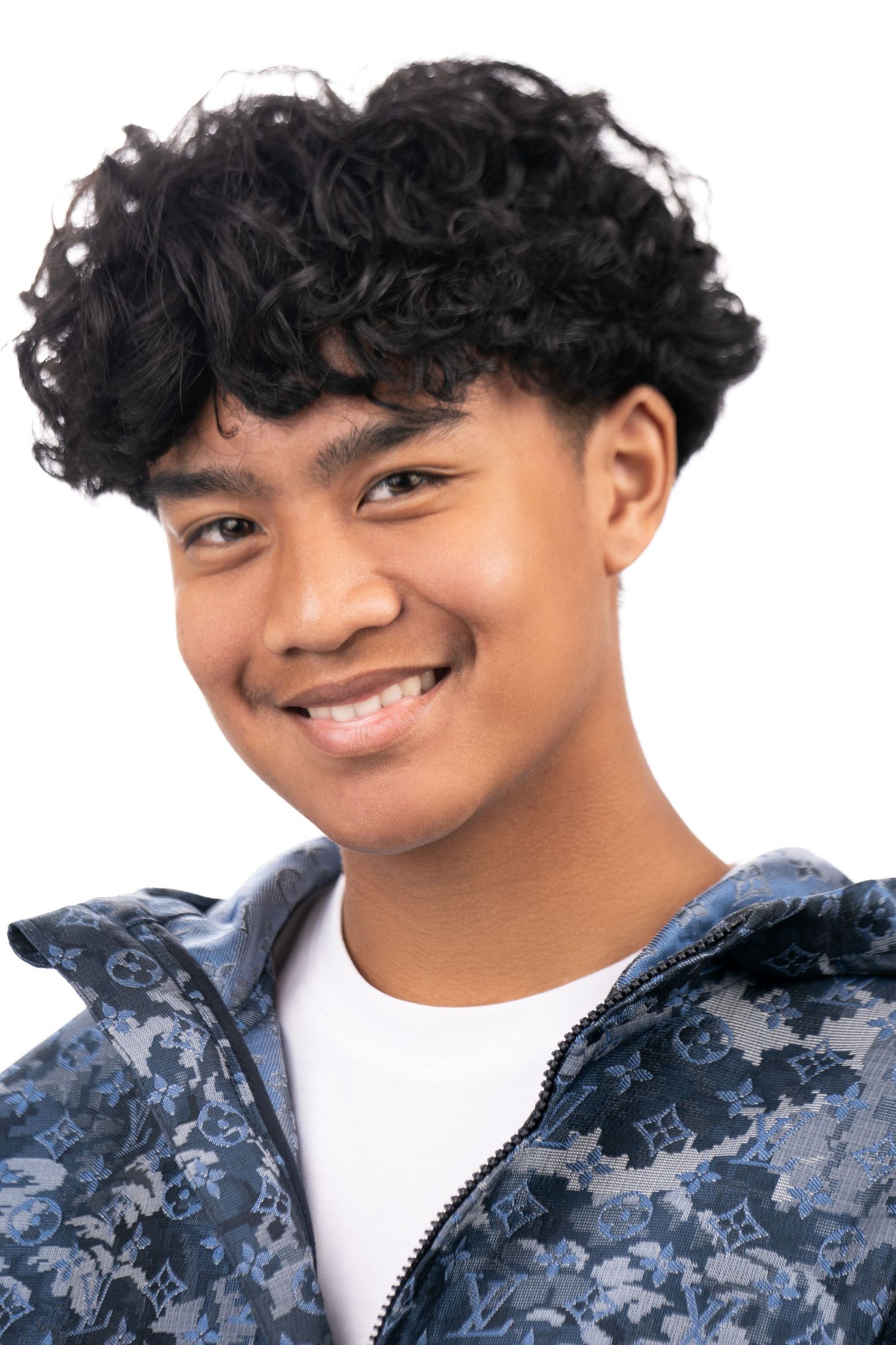 Portrait of a smiling young person with curly black hair wearing a white t-shirt and patterned blue jacket against white background.