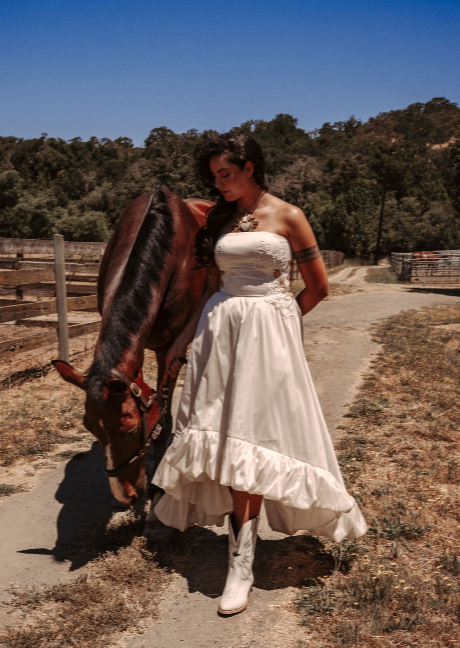 Woman in white wedding dress and cowboy boots stands next to brown horse in rustic ranch setting.