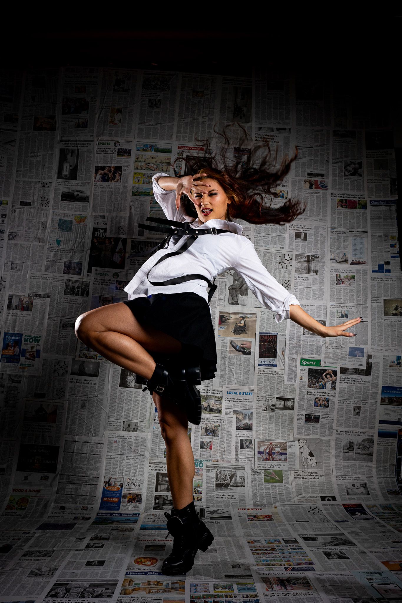 A dancer in a white shirt and black skirt leaps gracefully against a backdrop of newspaper pages in dramatic lighting.