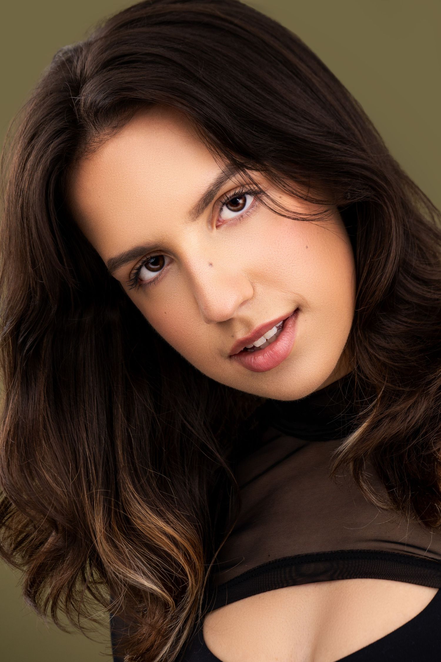 Beauty portrait with dark wavy hair wearing brown mesh top against olive background.