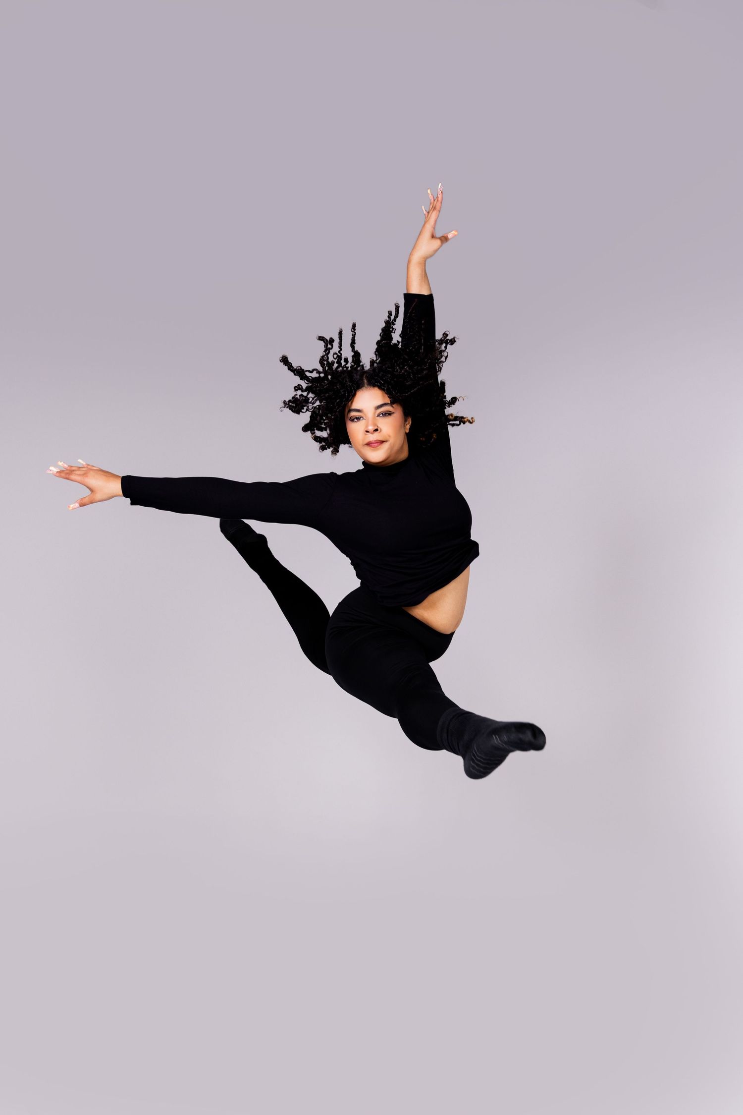 Dancer in black athletic wear performs a dramatic split leap against a gray studio background.