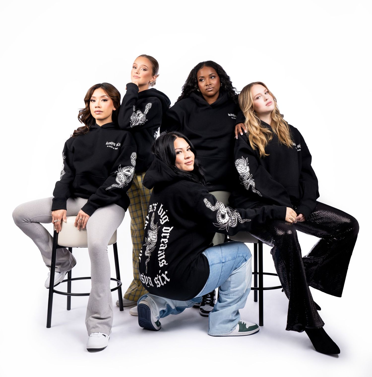 Group wearing coordinated black hoodies and casual bottoms seated on bar stools in studio setting.