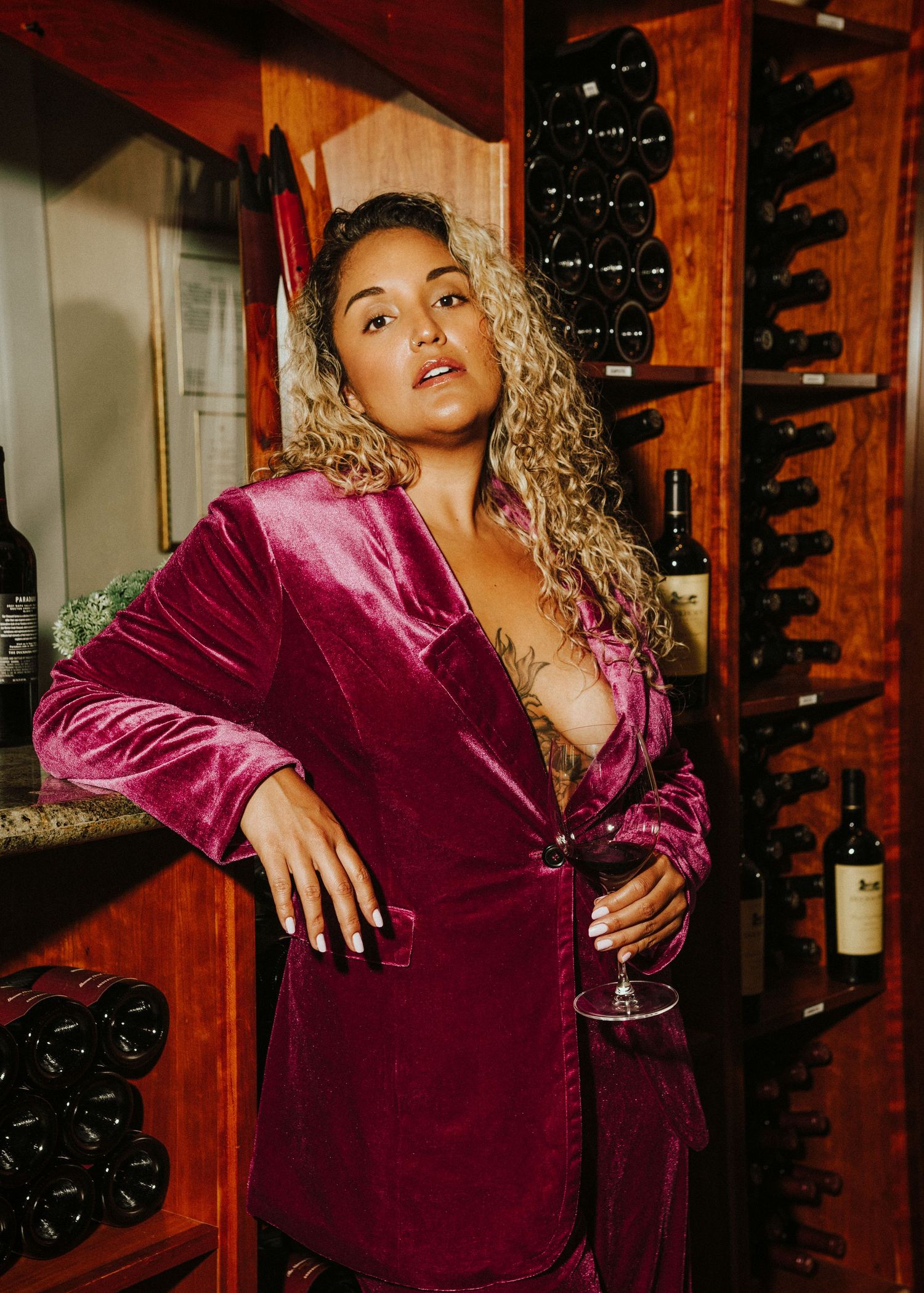 Fashion portrait in wine cellar with person wearing luxurious burgundy velvet blazer holding wine glass.