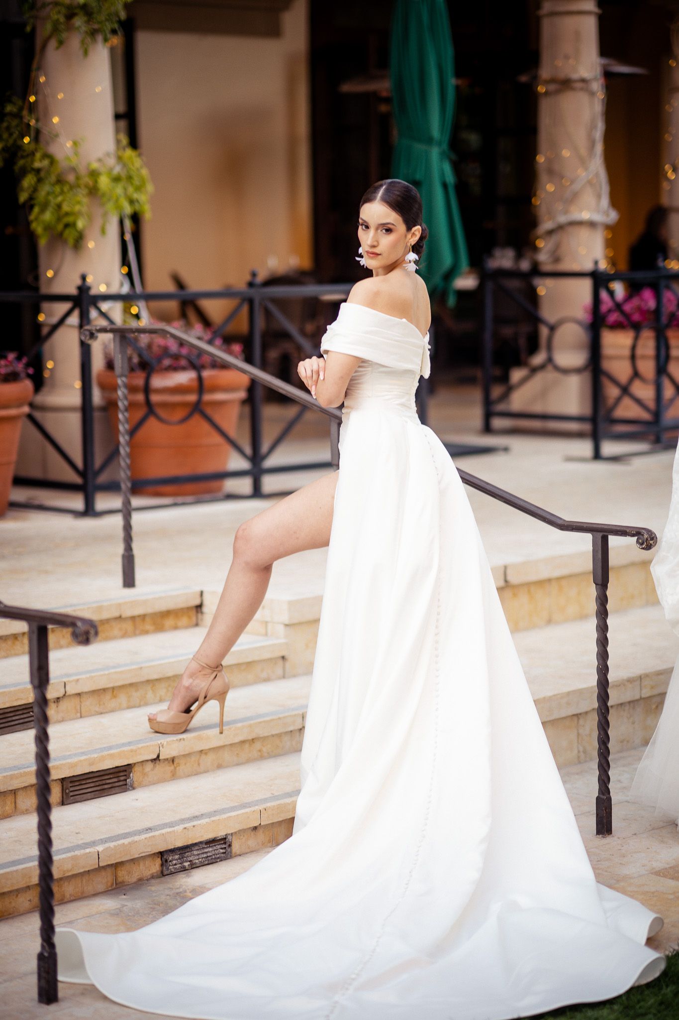 A person in a white off-shoulder wedding gown with a flowing train poses elegantly on stone steps with a wrought iron railing.
